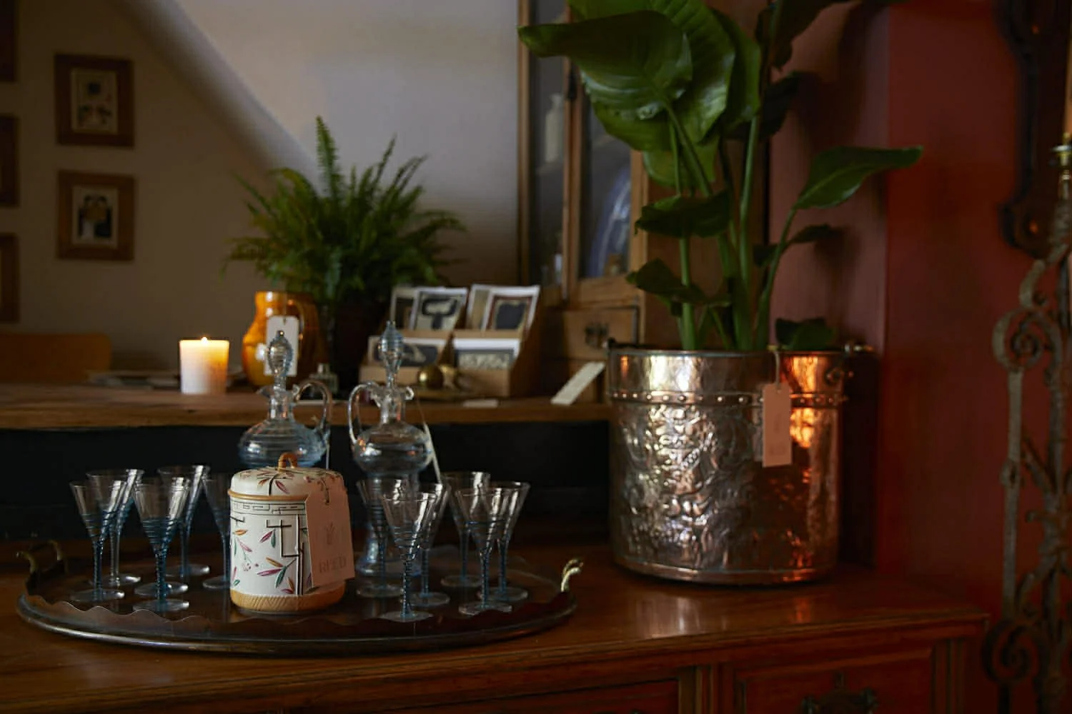 Decorative tabletop with glass decanters, glasses, potted plants, candles, and framed pictures in a cozy room.