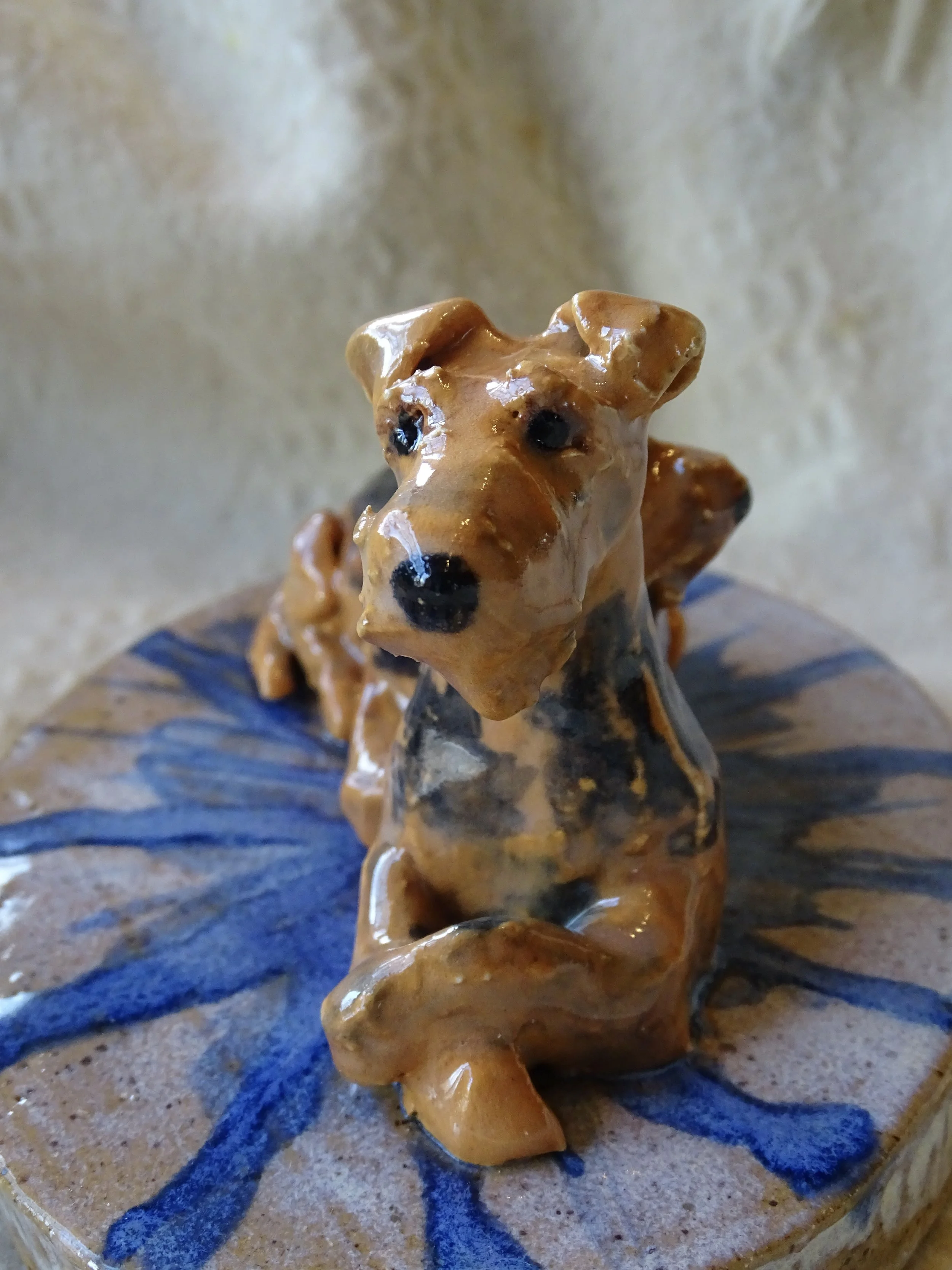 Ceramic sculpture of 2 Airedales sitting on a decorated base with blue and beige colors.