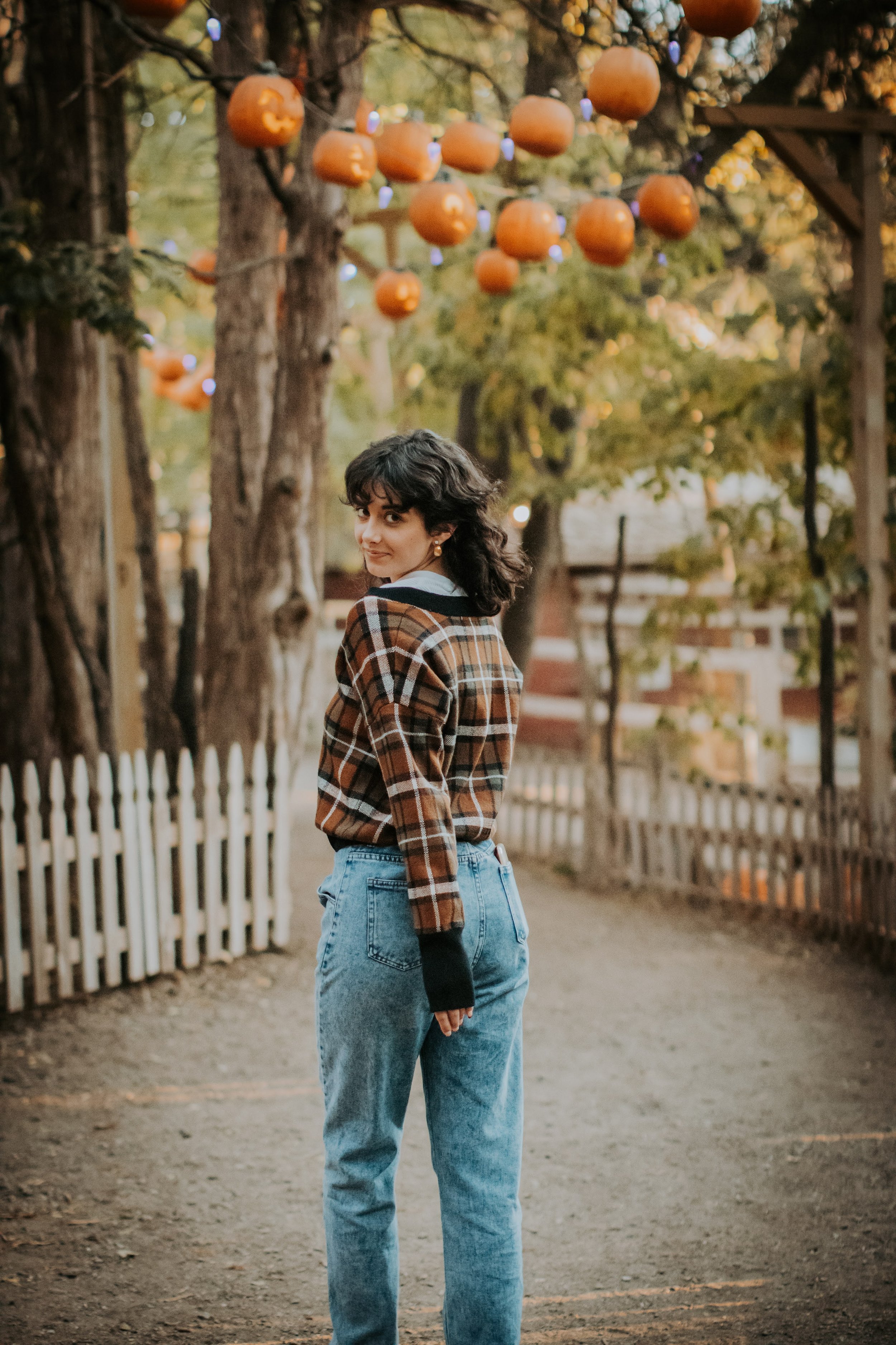 A young woman standing outdoors on a dirt path, with a white picket fence on one side and trees with hanging orange lanterns above. She is turned slightly, smiling and looking back, wearing a brown plaid shirt and high-waisted blue jeans.