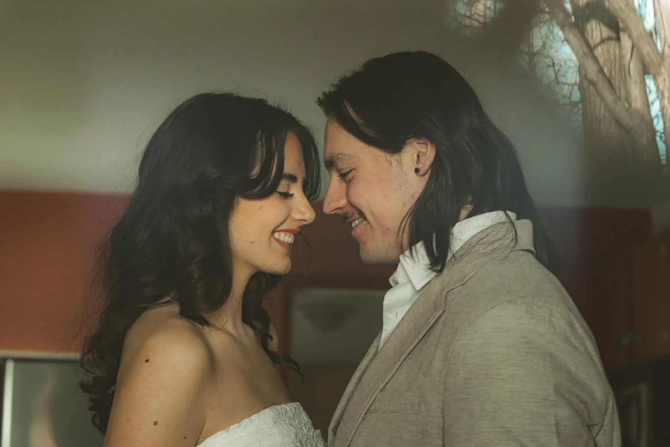 A woman and a man face each other closely, smiling with their foreheads almost touching, in a warm indoor setting.