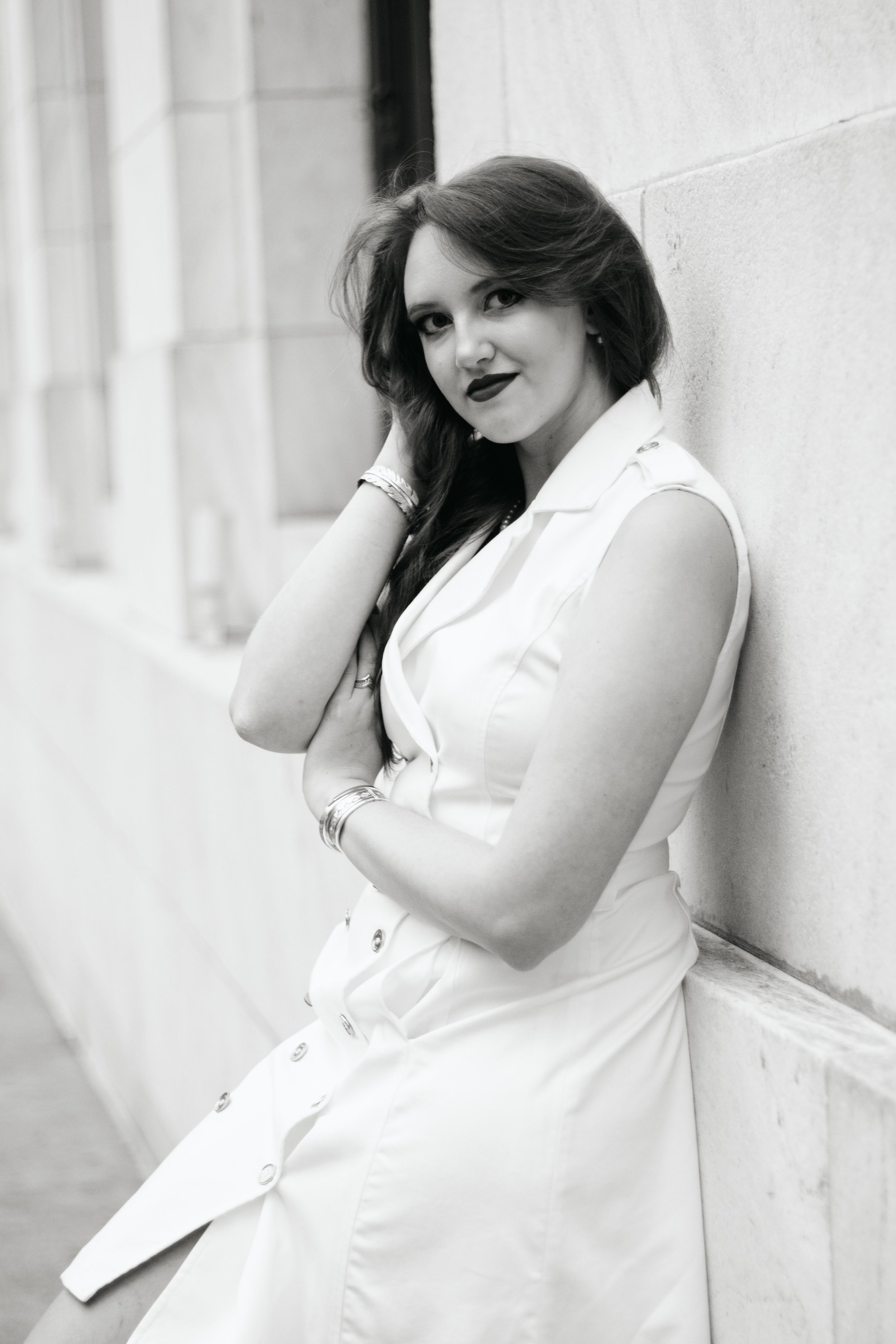 Black and white photo of a woman with long dark hair, wearing a sleeveless white dress, sitting and leaning against a stone wall, looking at the camera.