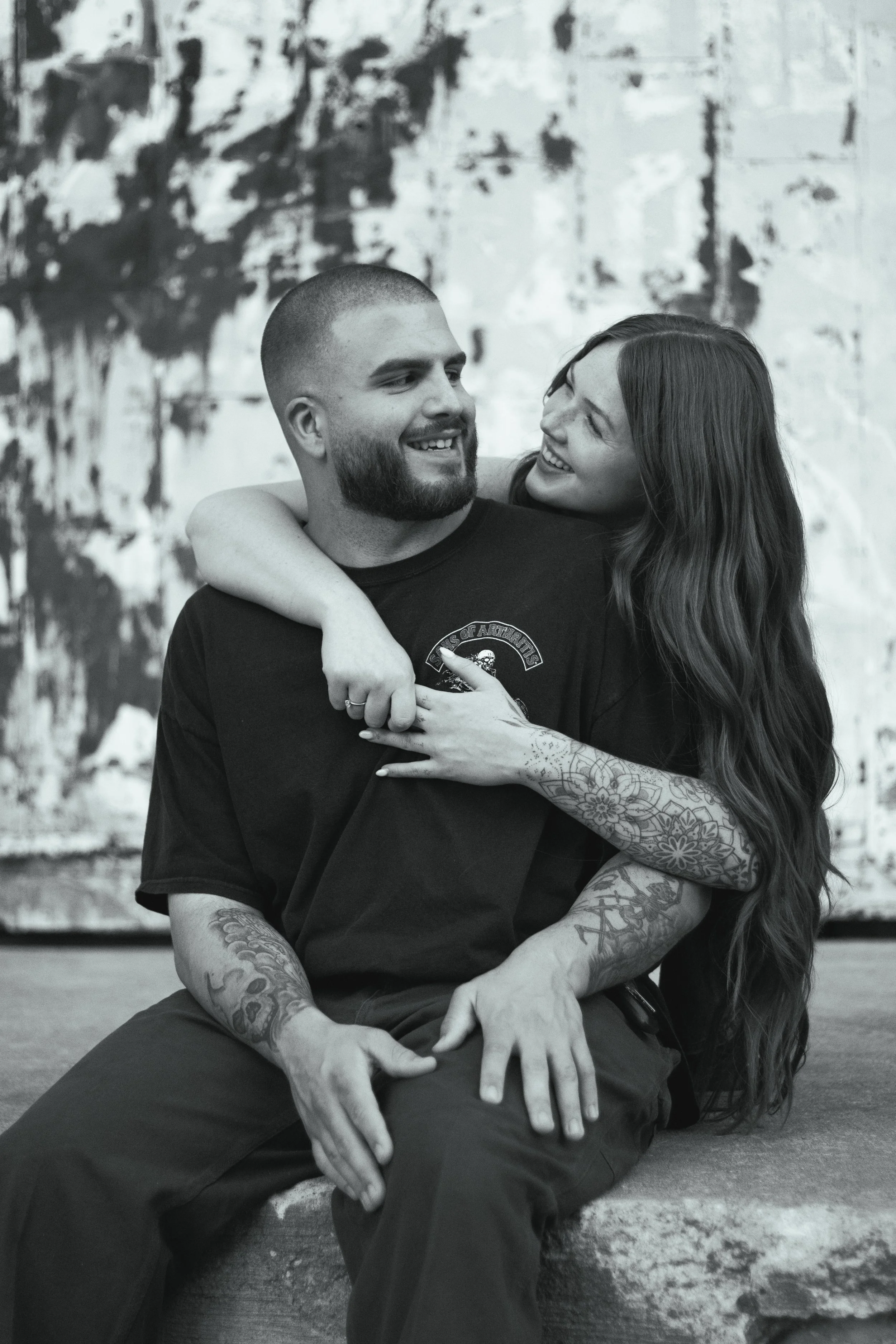A couple sitting on a ledge, smiling and embracing each other, with a painted wall in the background.