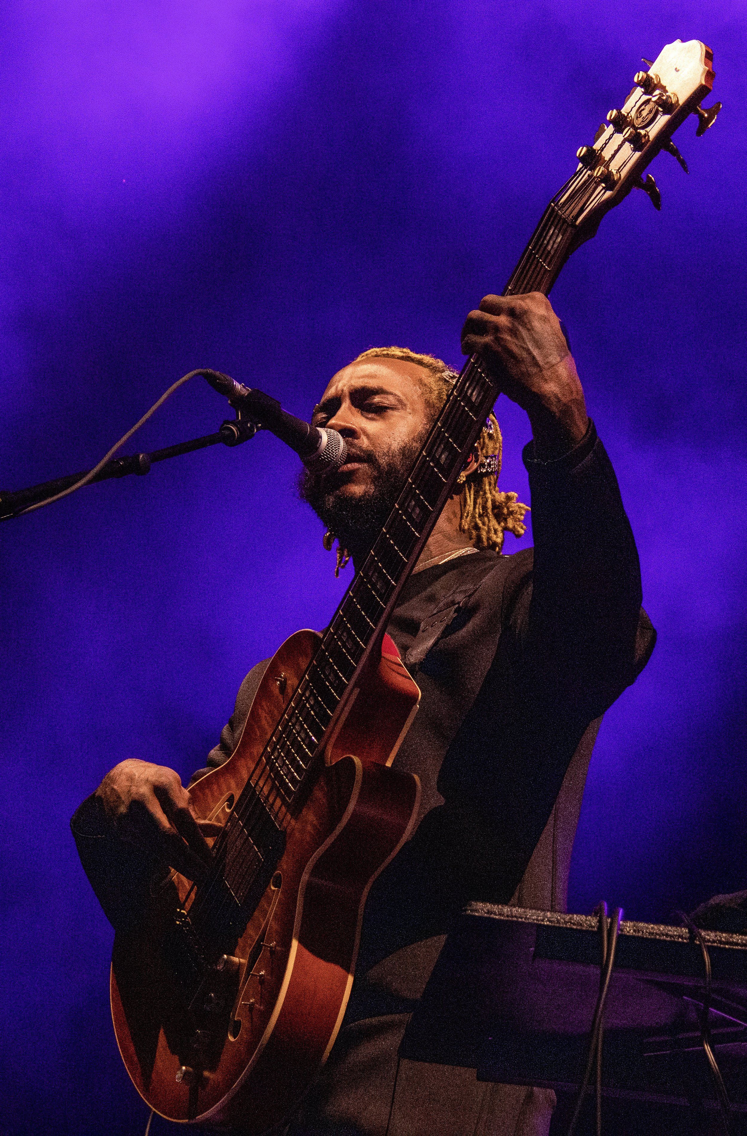 Musician with dreadlocks singing into a microphone while playing an acoustic guitar on stage with purple lighting.