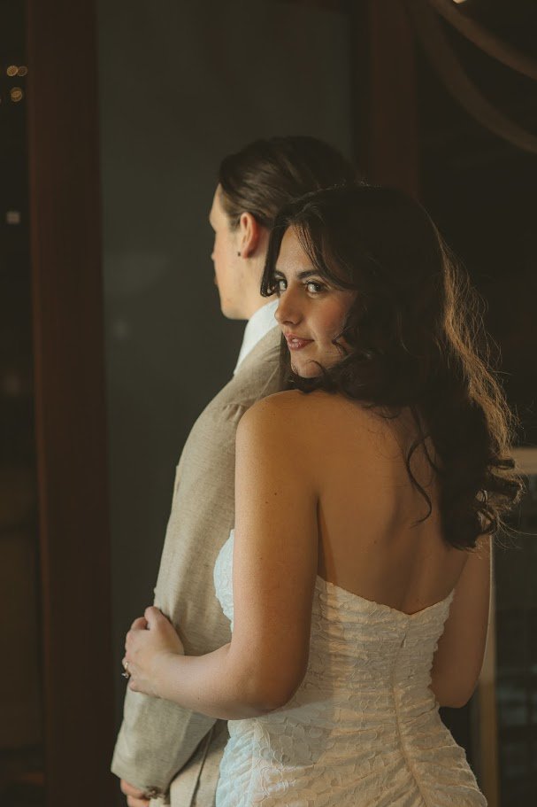 A woman in a strapless white dress with dark, curly hair smiling and looking at the camera, standing behind a man in a beige suit with dark hair. They are indoors near a mirror or glass surface.