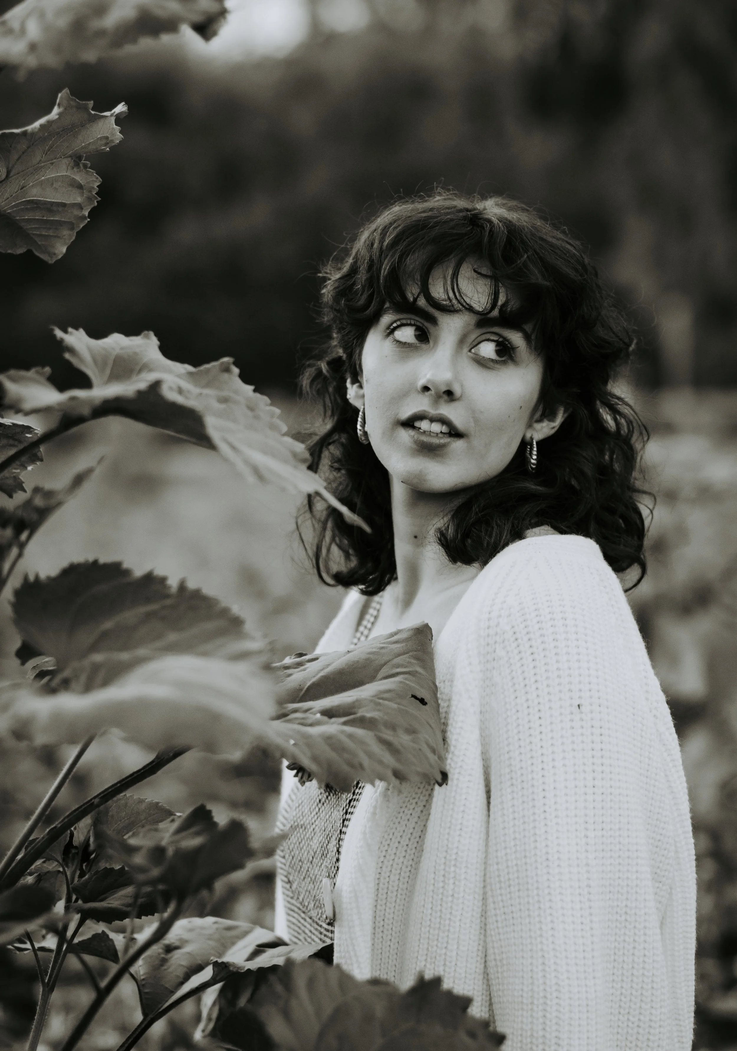 A black-and-white photo of a woman with curly hair, wearing earrings and a white sweater, standing among large leaves outdoors.