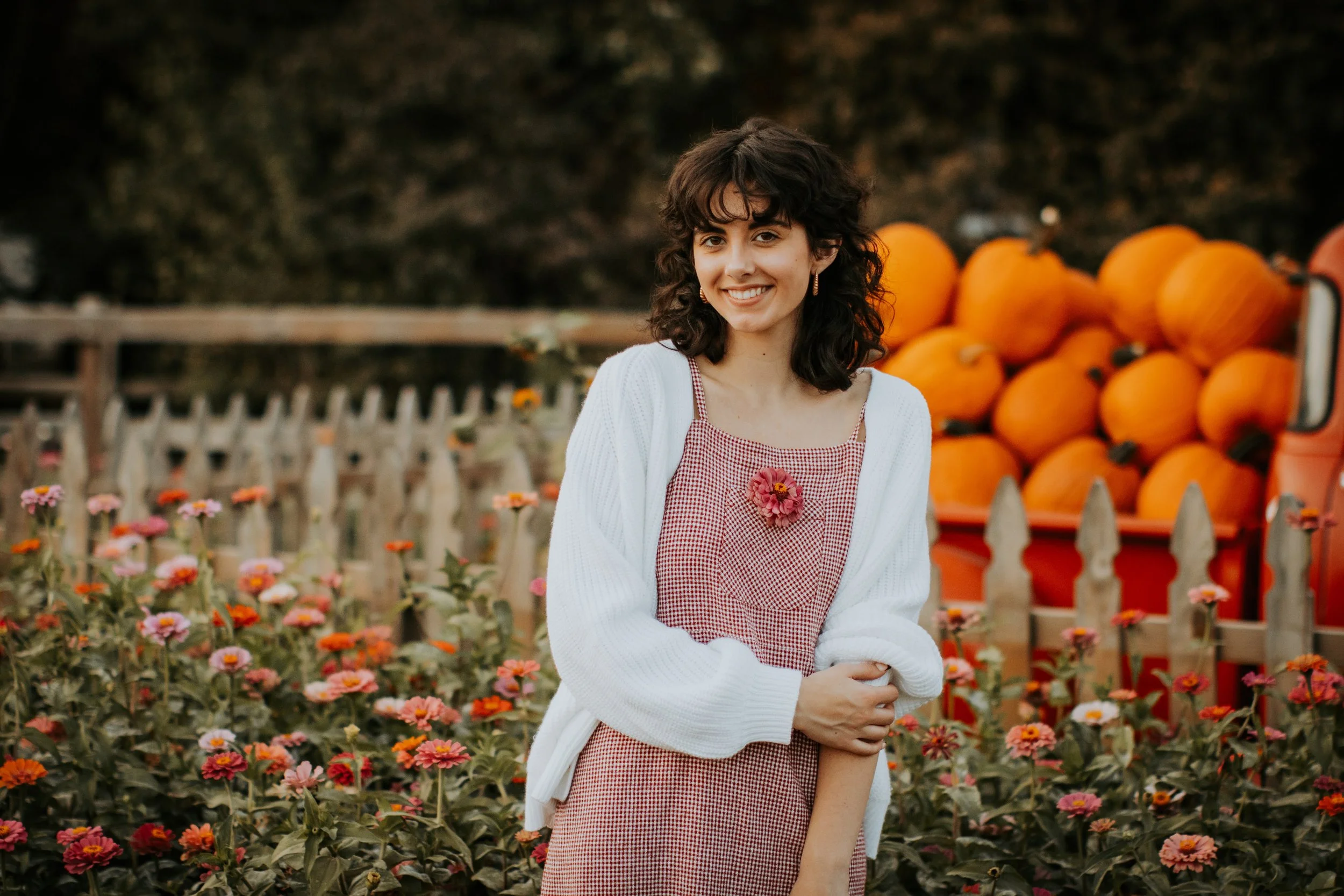 A young woman with dark curly hair smiling outdoors at a pumpkin patch surrounded by orange pumpkins and pink flowers, wearing a checked dress and white cardigan.