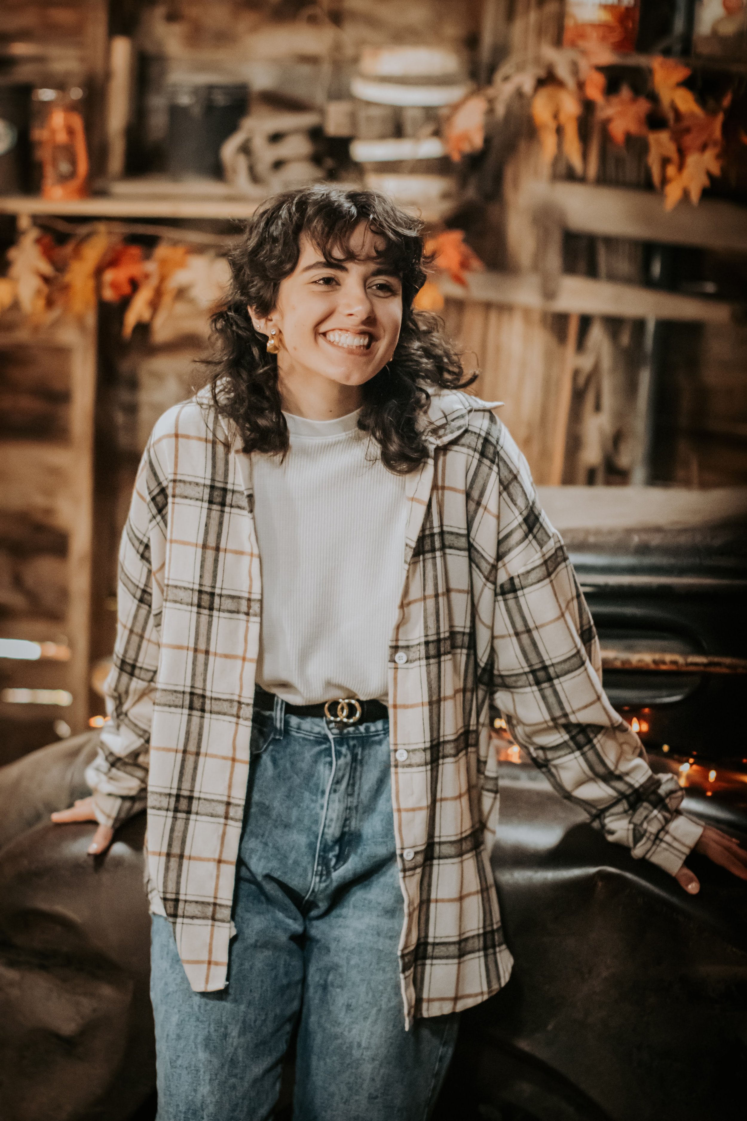 A young woman with curly dark hair smiling, standing indoors with a wooden background decorated with autumn leaves, wearing a white turtleneck, a beige plaid shirt, and high-waisted blue jeans.