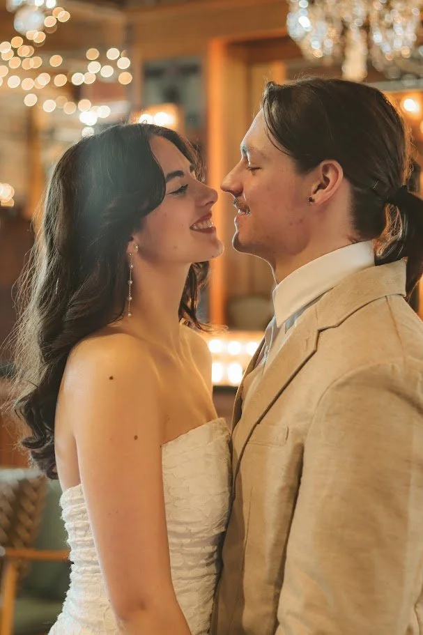A couple standing close together indoors, smiling and about to kiss in a warmly lit room with string lights and chandeliers.