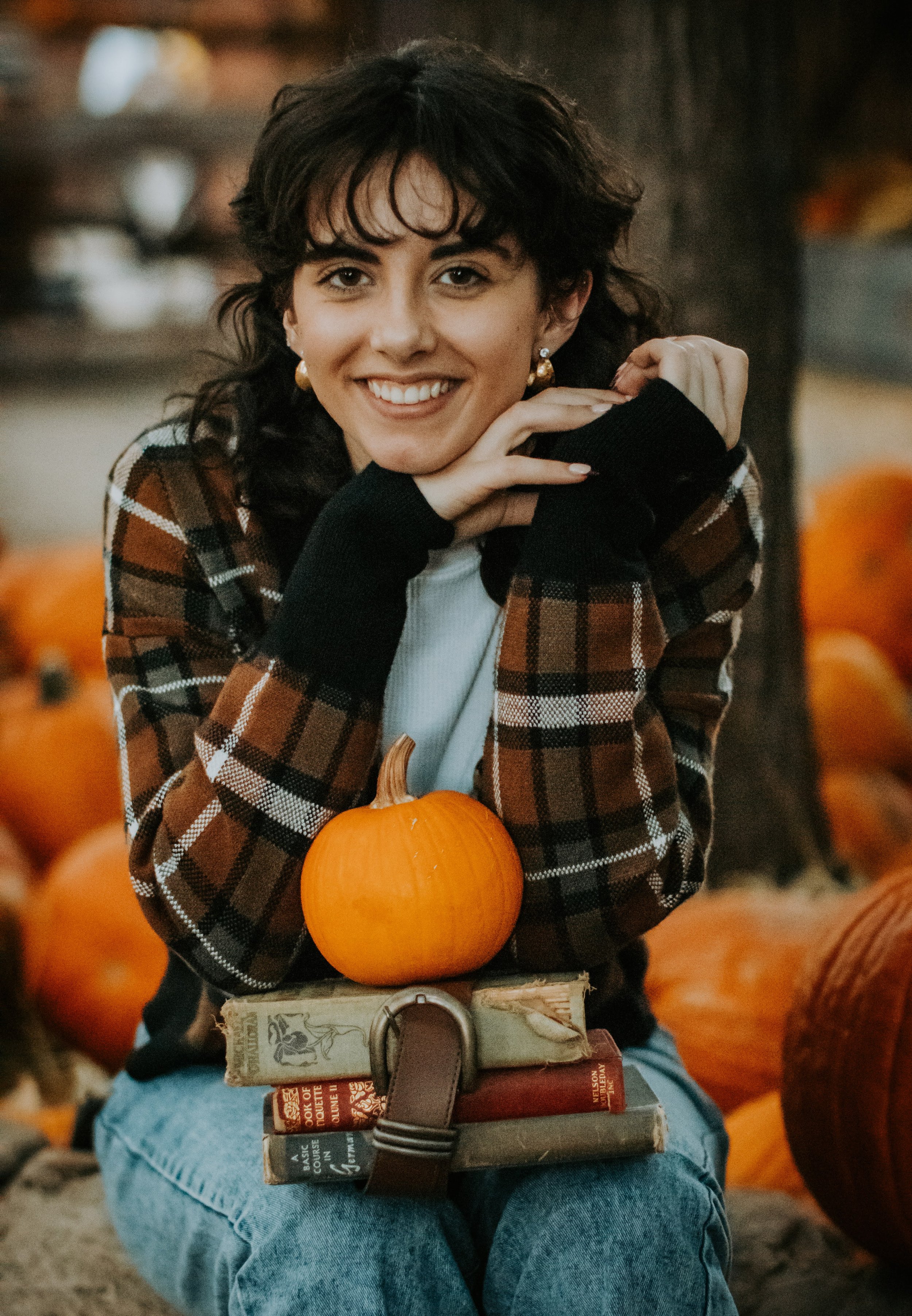 A woman smiling at a pumpkin patch, wearing a plaid jacket, surrounded by pumpkins, with a pumpkin resting on her lap atop books and a belt.