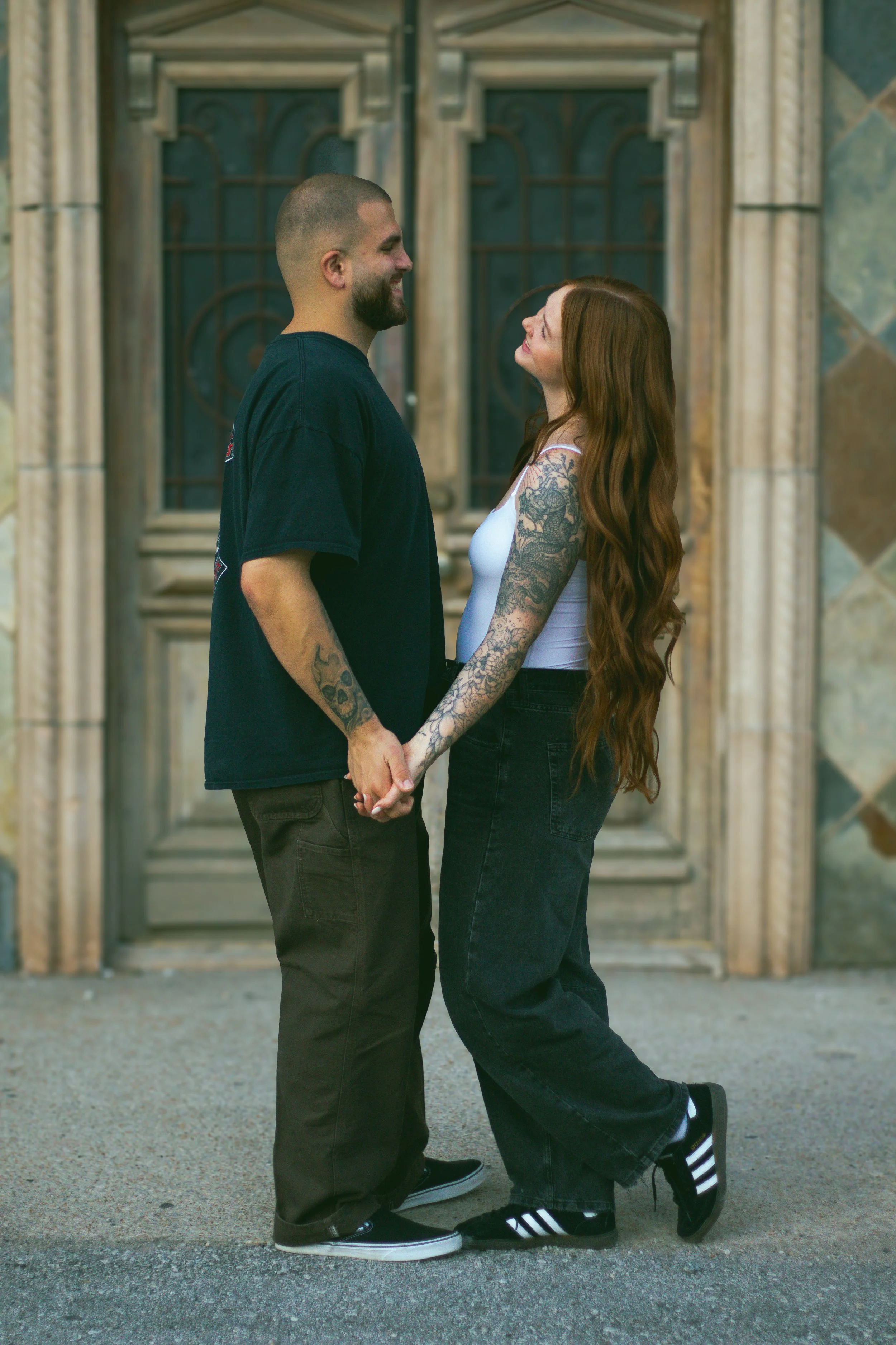 A couple holding hands and smiling at each other in front of a decorative wooden door.
