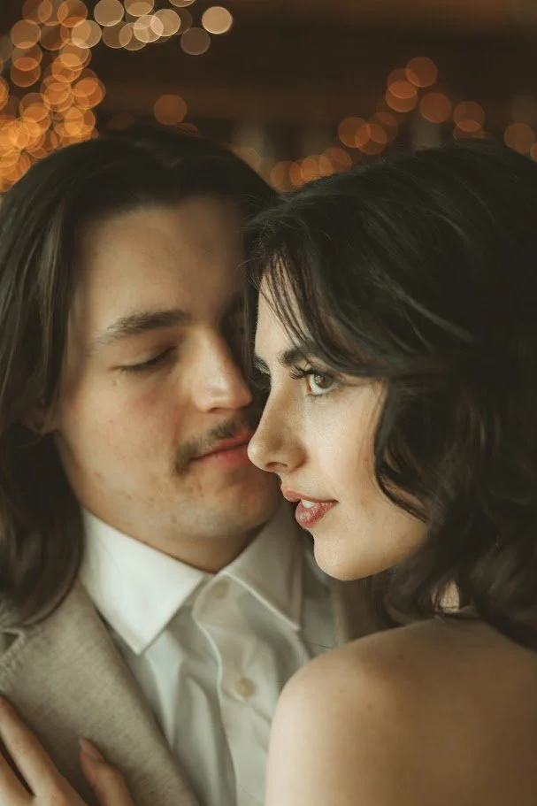 A close-up of a romantic moment between a man with long hair and a woman with black hair, with blurred string lights in the background.