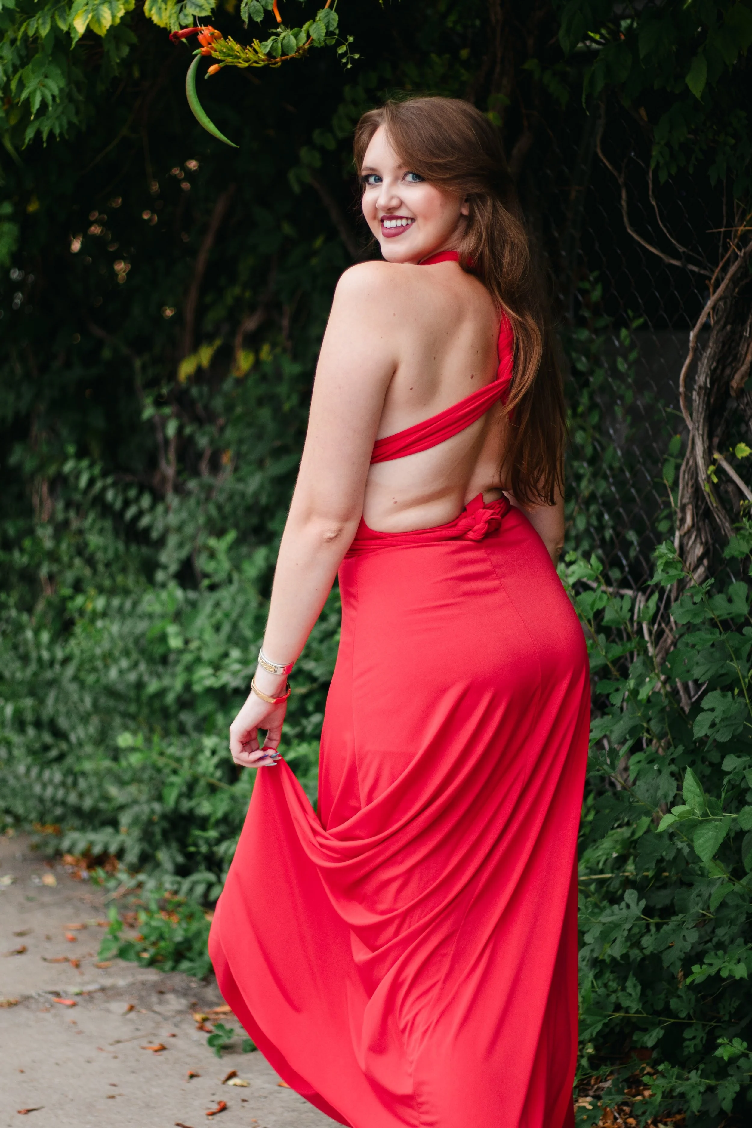A woman in a red dress with a backless and open side design, standing outdoors near greenery.