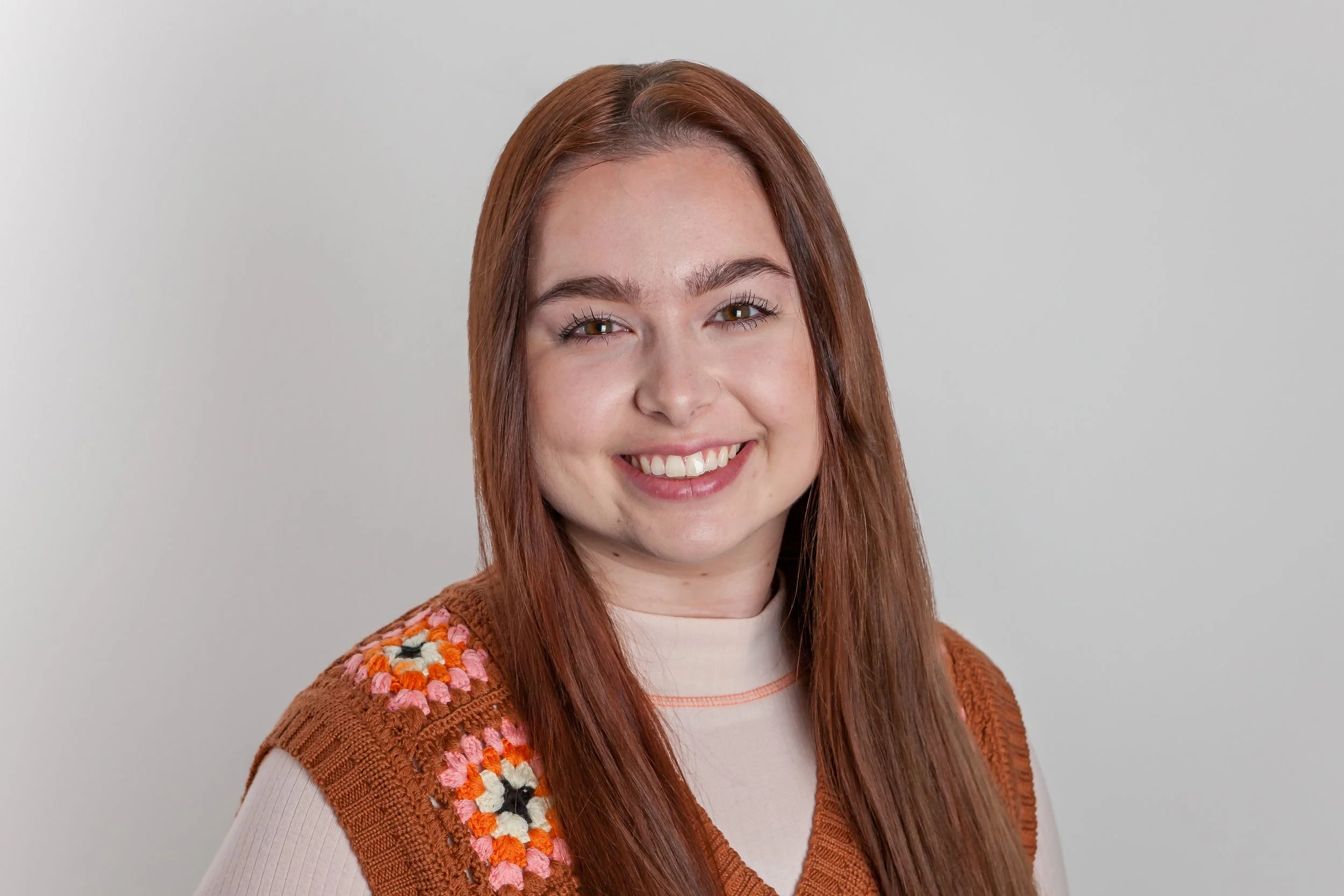 A young woman with long reddish-brown hair, smiling, wearing a beige top and a brown knitted vest with colorful floral patterns on the shoulder, standing against a plain gray background.