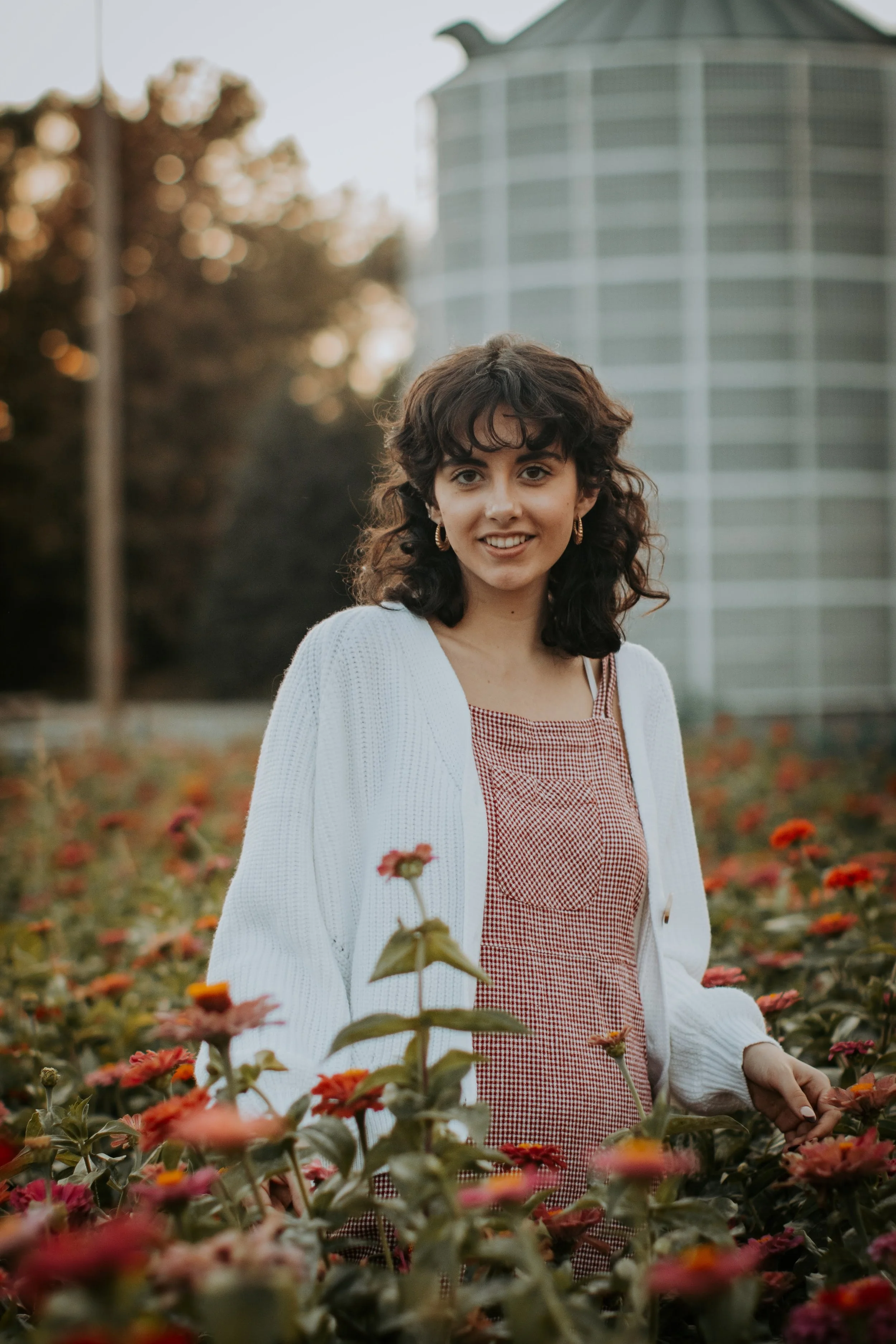 A woman with curly dark hair smiling at the camera in a field of orange and pink flowers, with a large building in the background.