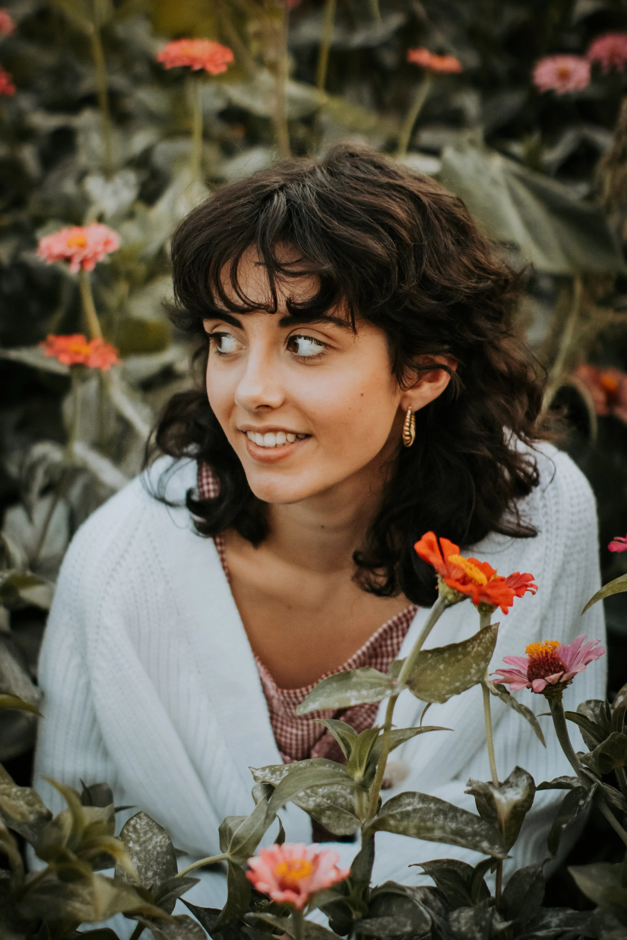 A young woman with dark, curly hair and hoop earrings smiling, surrounded by pink and orange flowers in a garden or flower field.