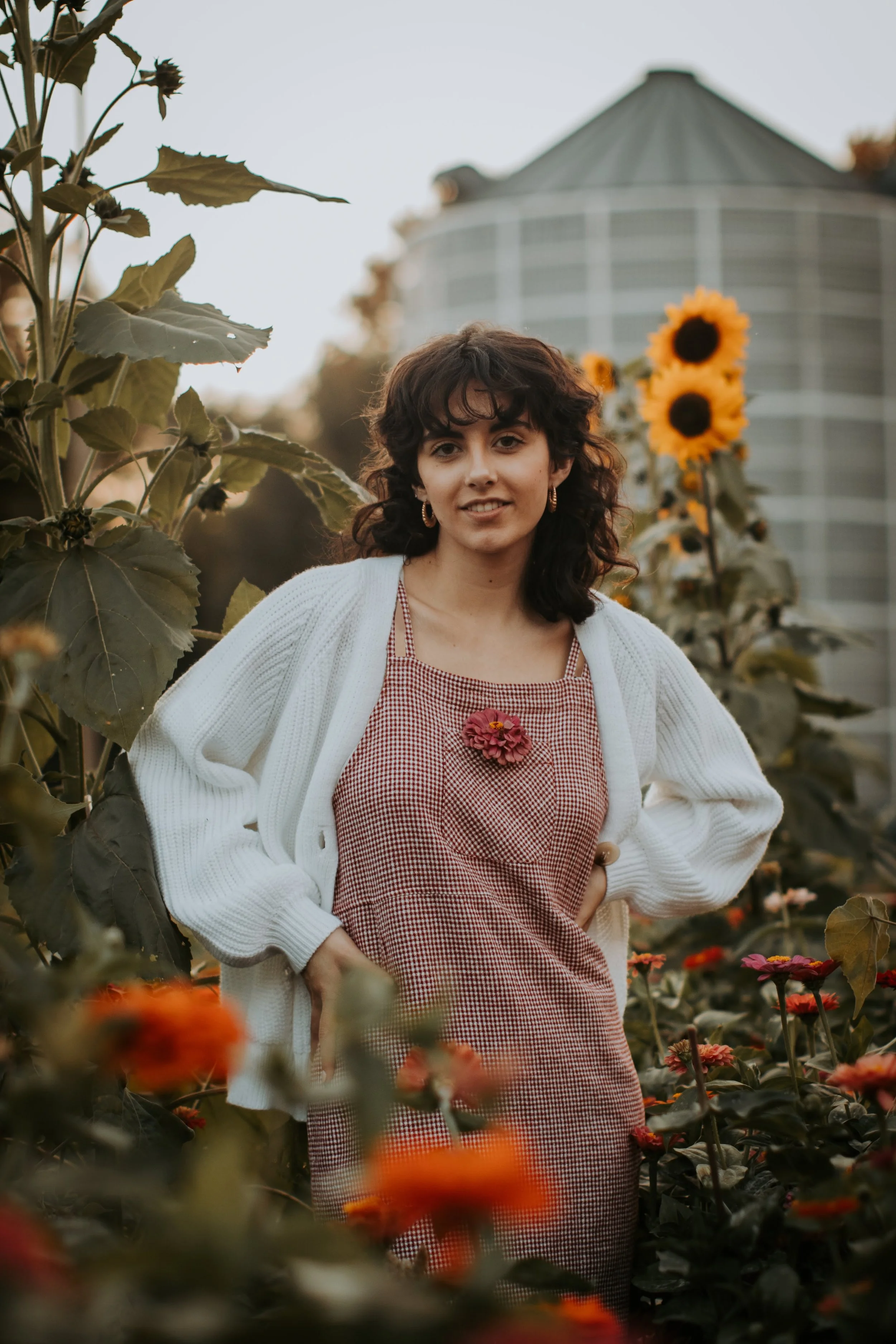 A young woman with curly dark hair and hoop earrings standing in a flower garden with sunflowers and other vibrant flowers, with a greenhouse in the background during sunset.