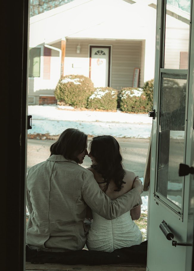 A couple sitting on a porch or step outside a house, embracing each other and smiling, with snow on the ground and bushes in the background.
