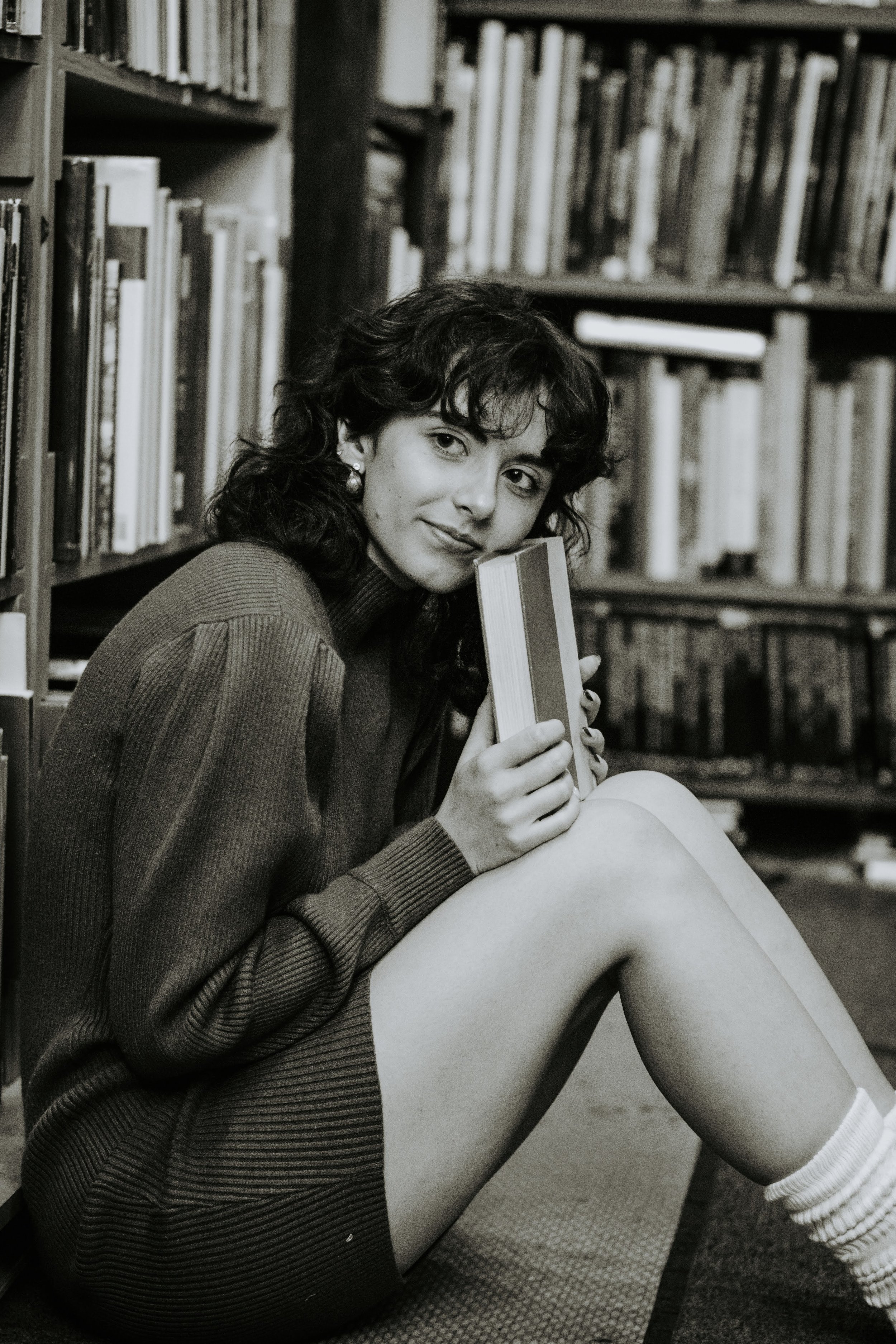 A young woman with curly dark hair sitting on the floor of a library, holding a book close to her chin, with shelves full of books behind her.