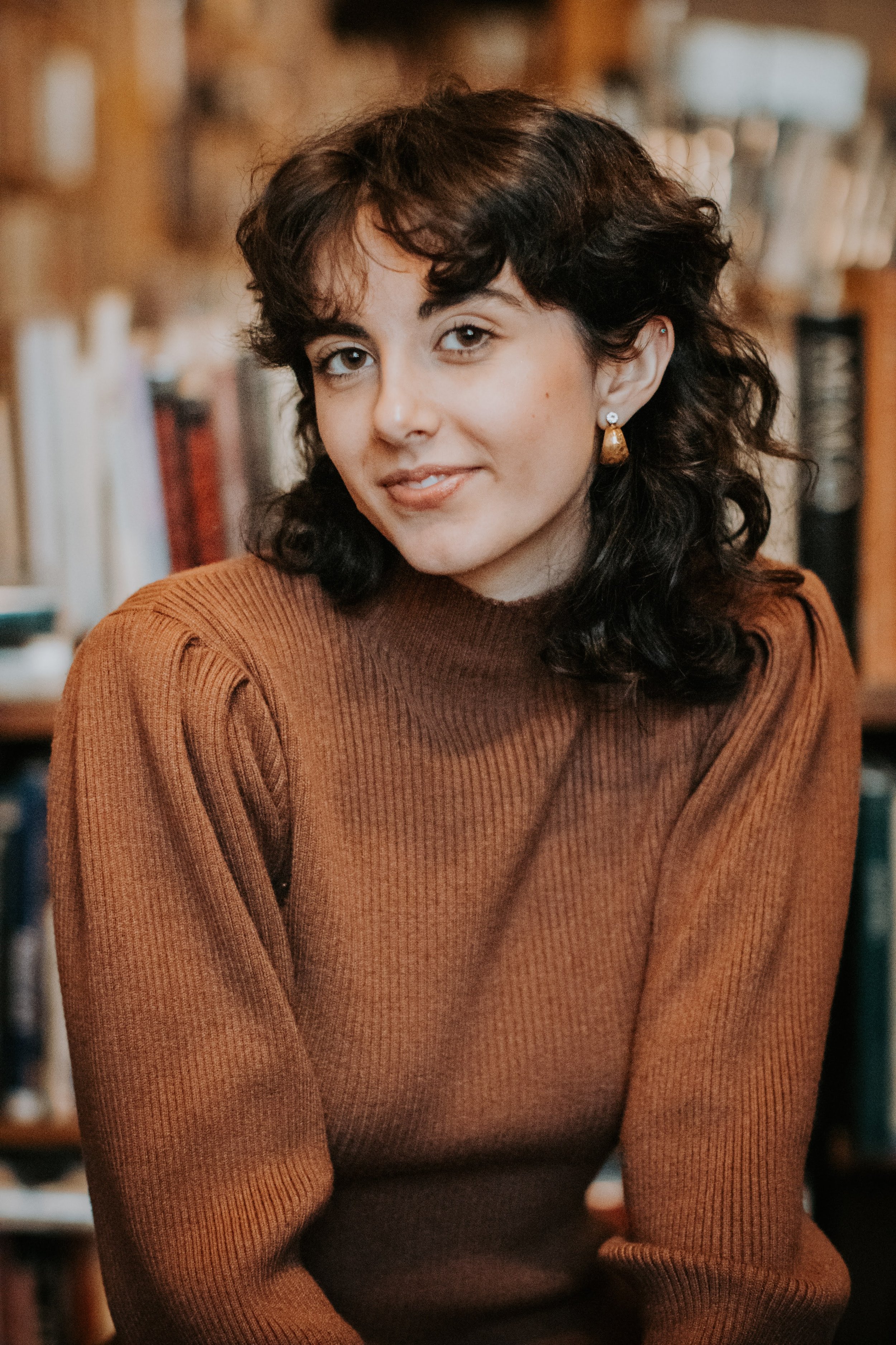 A young woman with dark curly hair wearing a brown ribbed sweater, sitting in a cozy room with a bookshelf in the background.