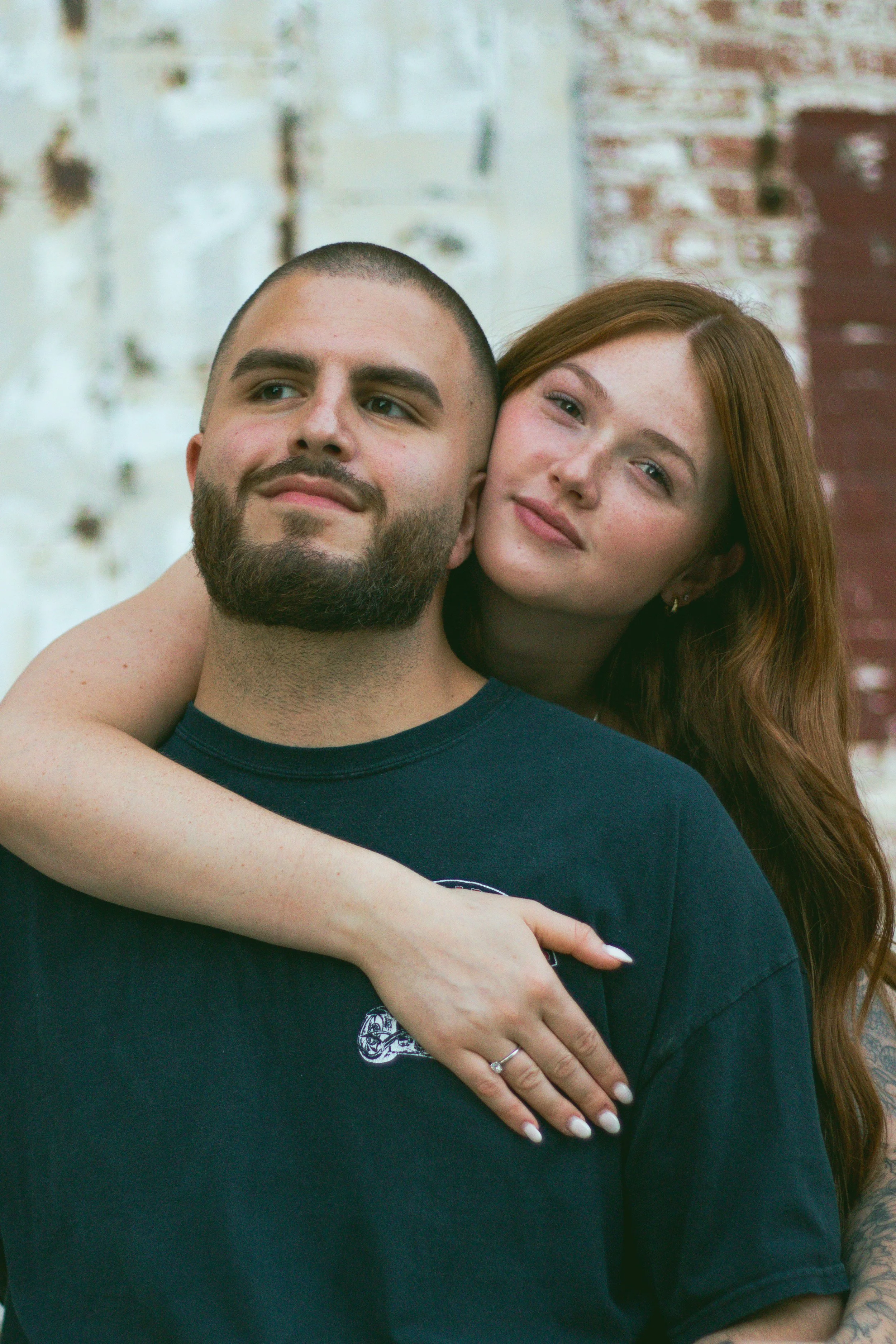 A close-up of a couple outdoors, with the woman embracing the man's shoulders and resting her head on his. They are standing in front of a brick wall.