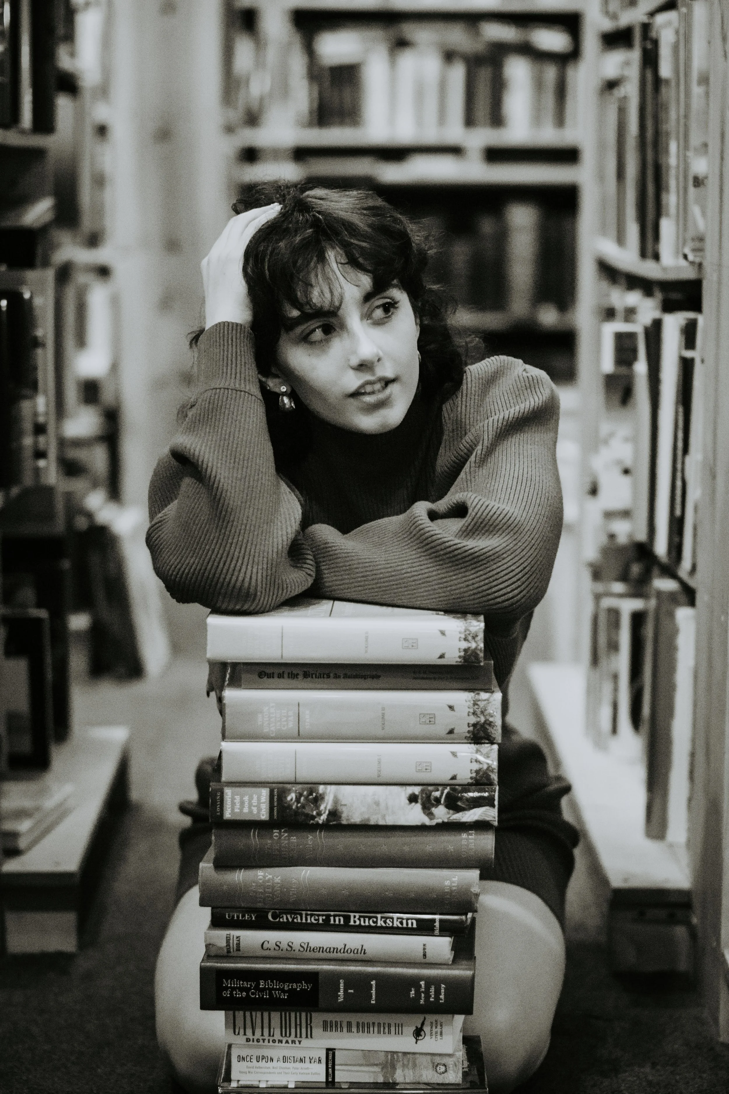 A young woman with dark hair and a sweater, sitting in a library, resting her head on her hand, leaning on a stack of books.
