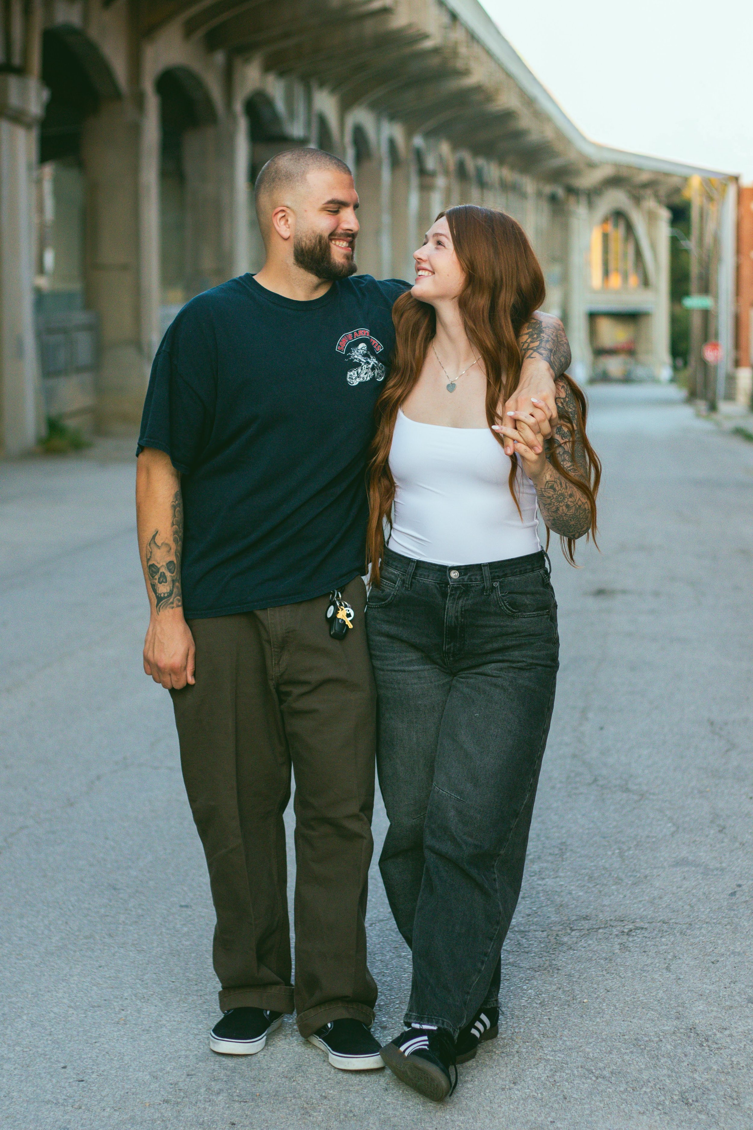 A young couple walking arm in arm on a street, smiling and looking at each other.