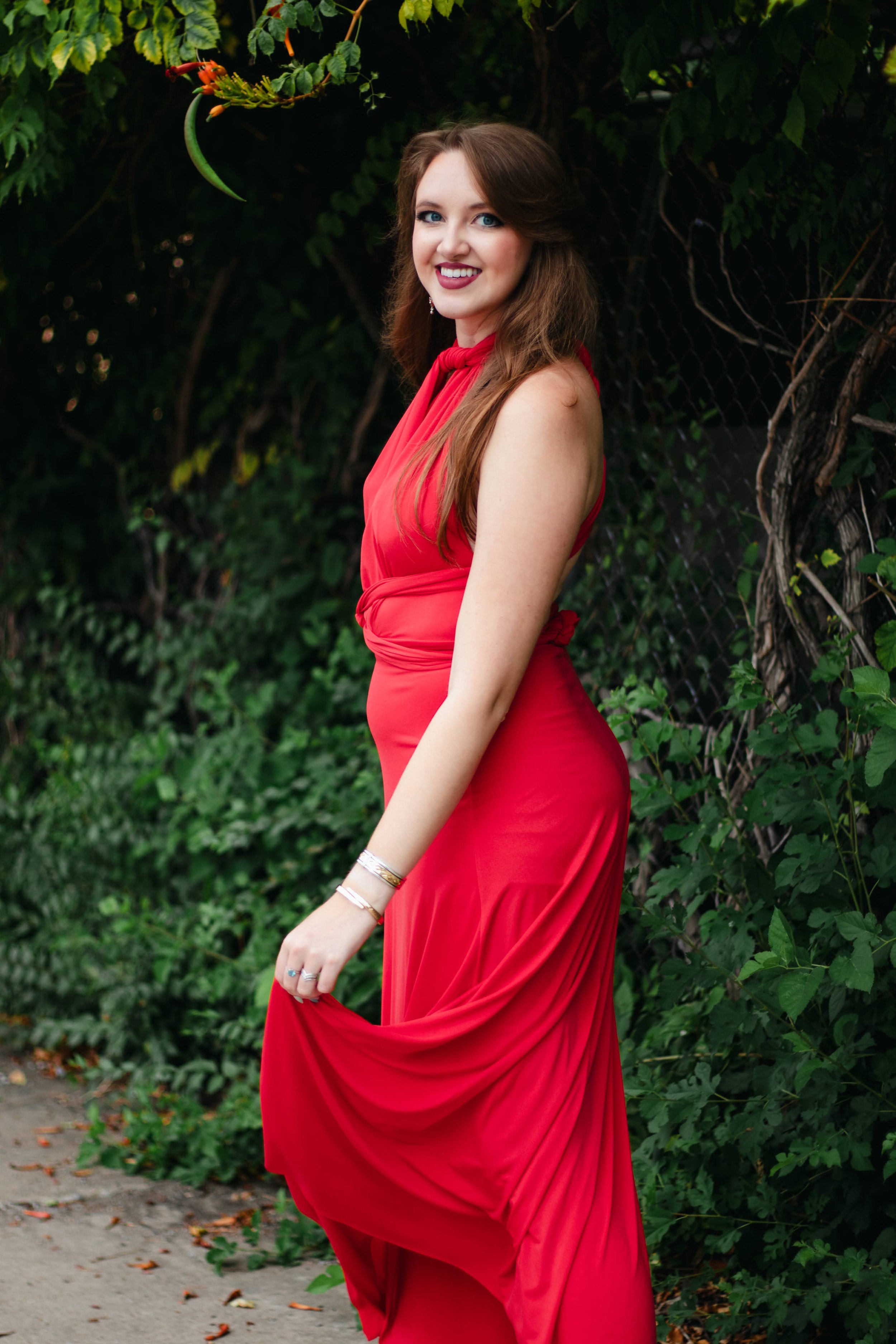 A woman in a red evening gown stands outdoors in front of green foliage, smiling at the camera.