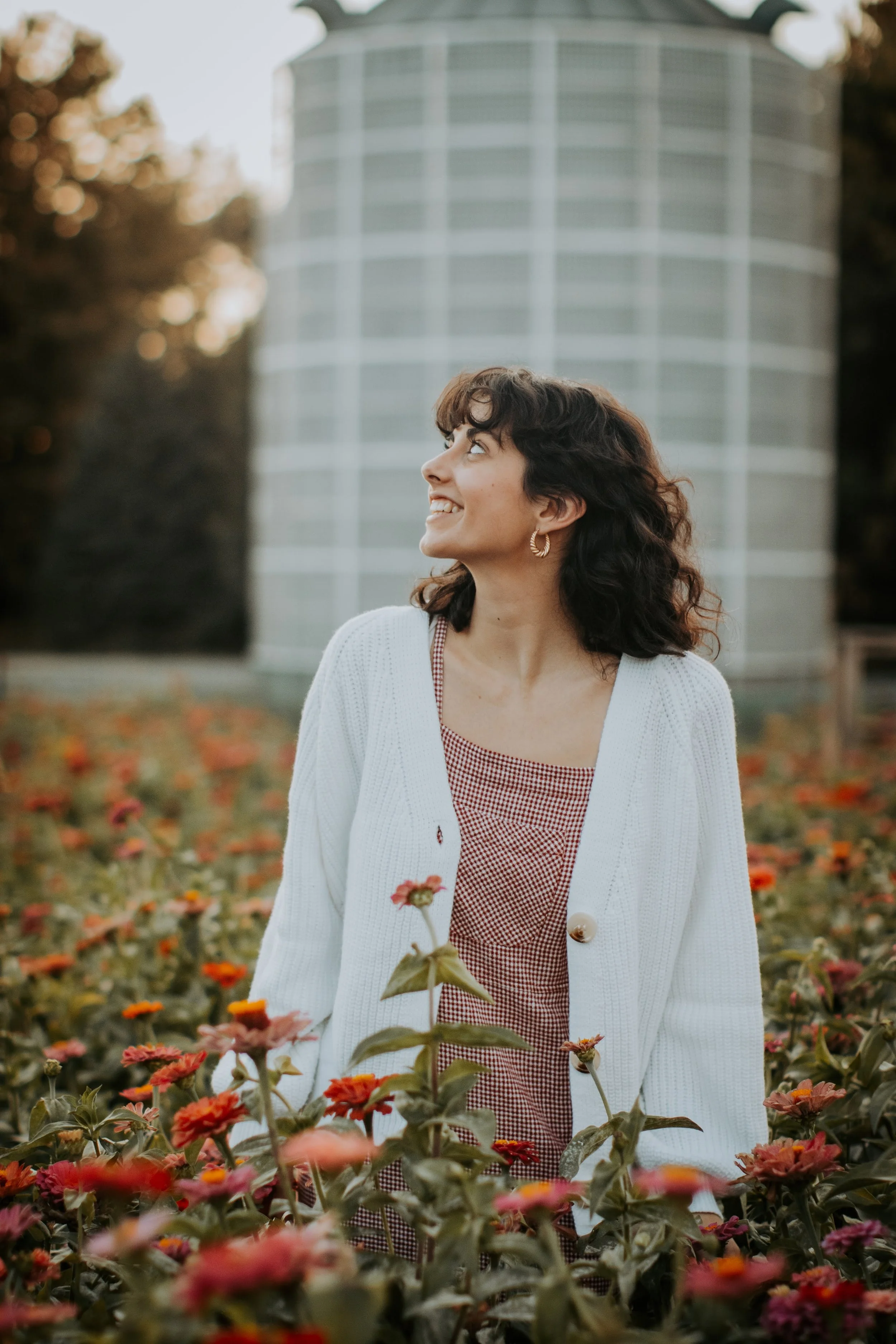 A woman with dark curly hair wearing a white cardigan and a red checkered dress, standing in a flower field and looking upward with a smile.