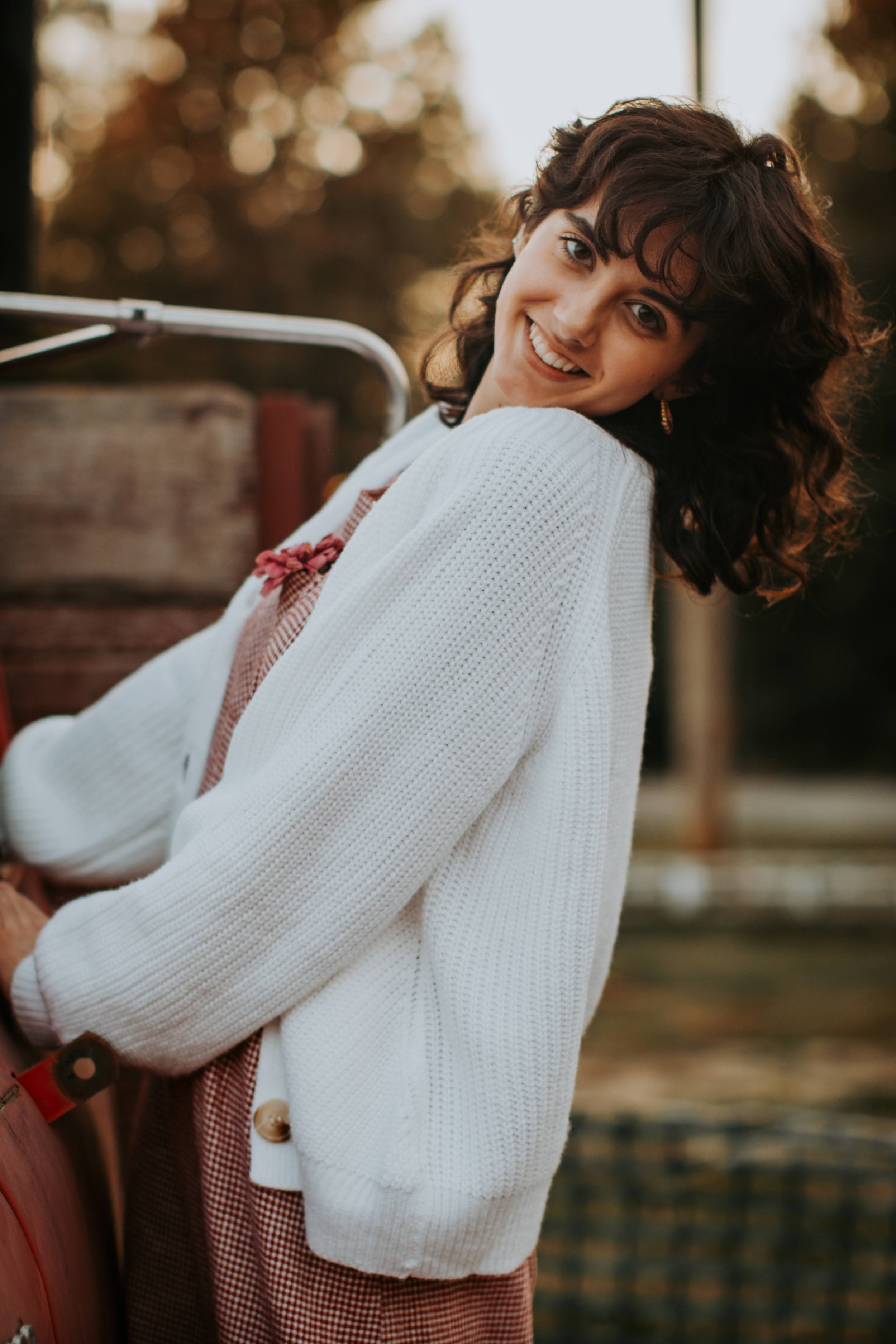 A woman with dark curly hair smiling outdoors during golden hour, wearing a white cardigan and brown checkered pants.
