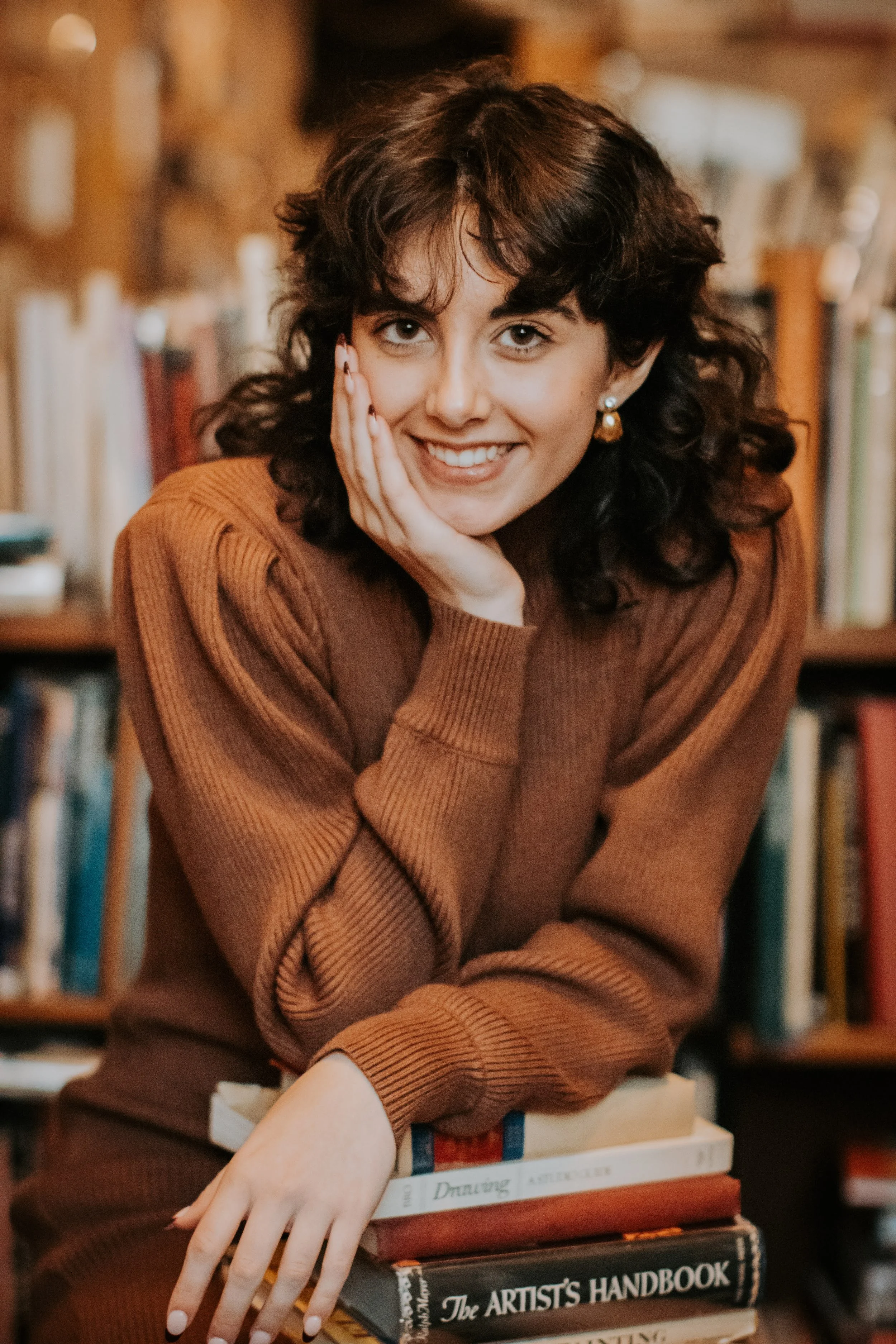 A young woman with curly dark hair, smiling, resting her face on her hand, sitting behind a stack of books in a cozy library setting.