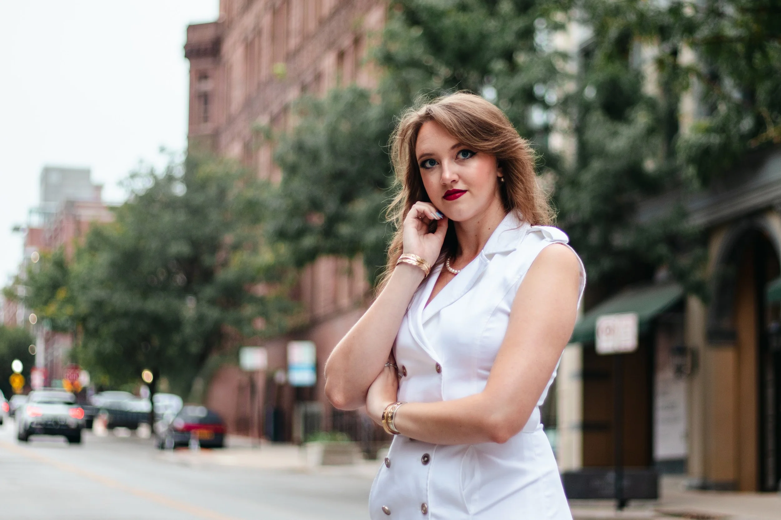A woman with light brown hair and red lipstick wearing a sleeveless white dress, standing on a city street with trees and brick buildings in the background.