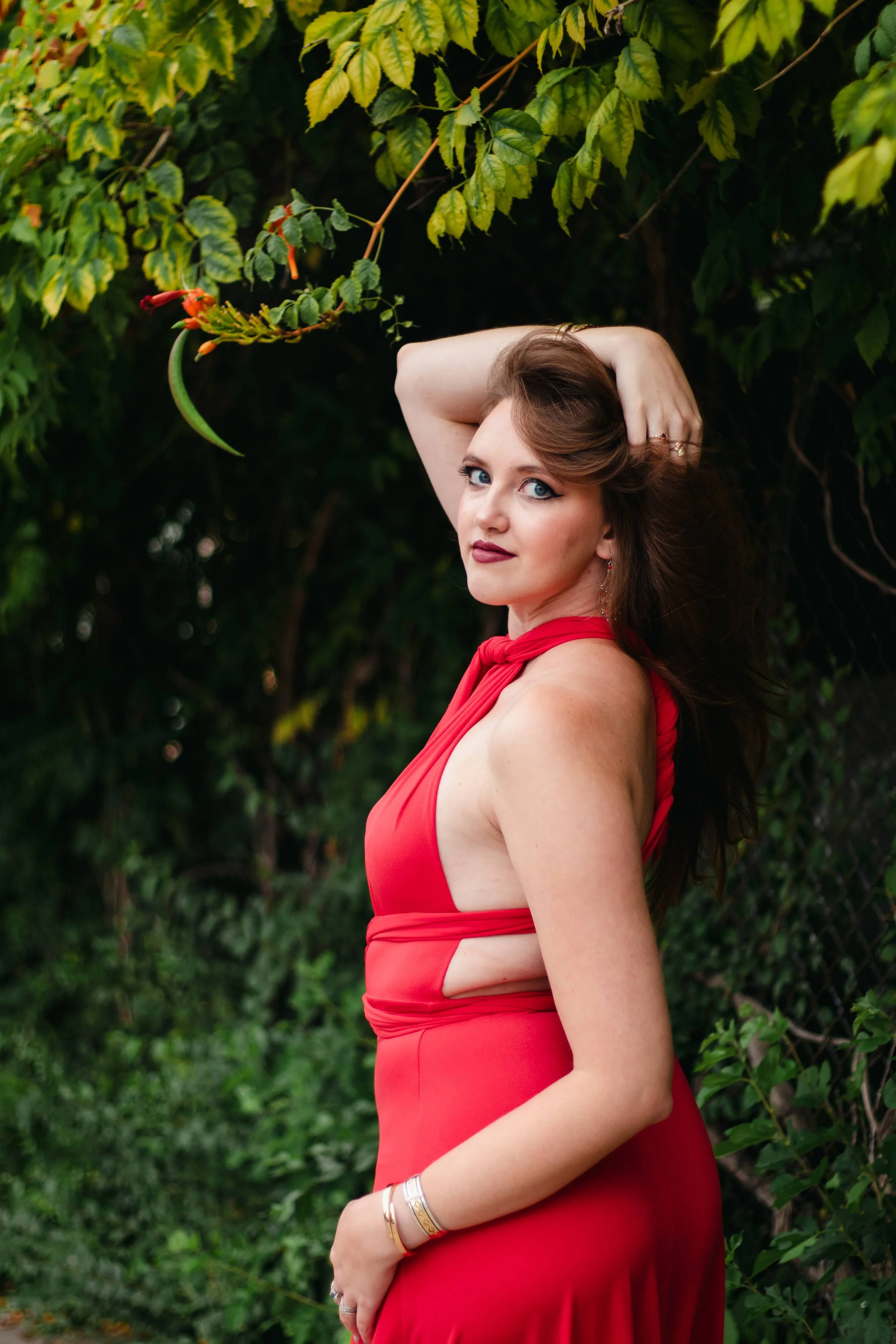 A woman with brown hair and blue eyes wearing a red dress with cutouts, standing outdoors in front of greenery, with her hand resting on her head.