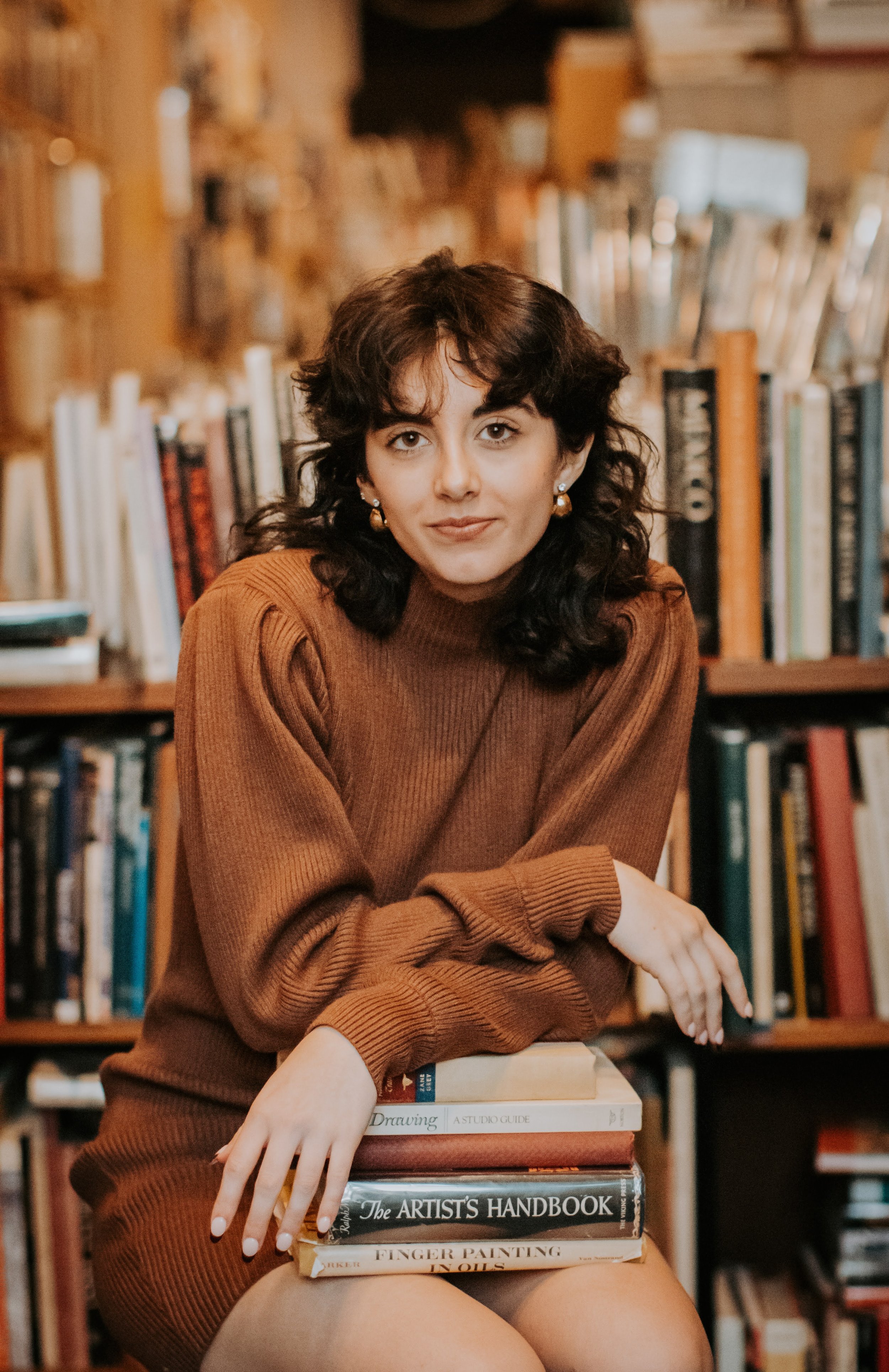 A woman with dark, curly hair and earrings, wearing a brown sweater, sitting in a library with books on shelves behind her, resting her arms on a stack of books.