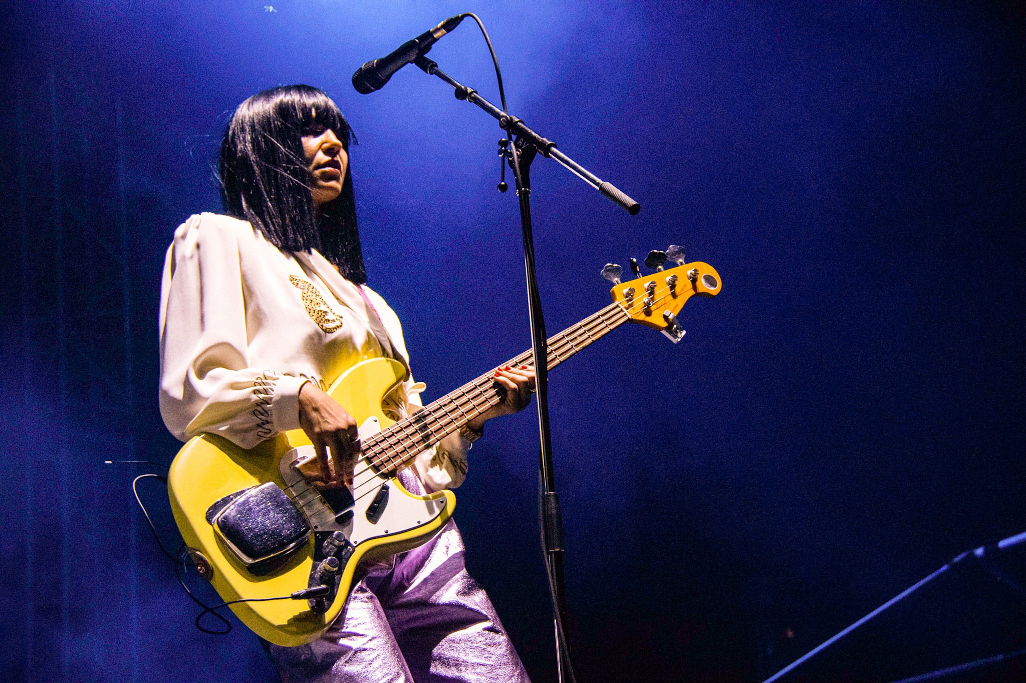 A woman with black hair and bangs, wearing a white blouse with leopard print details, playing a yellow bass guitar on stage with a microphone in front of her, against a dark blue background.