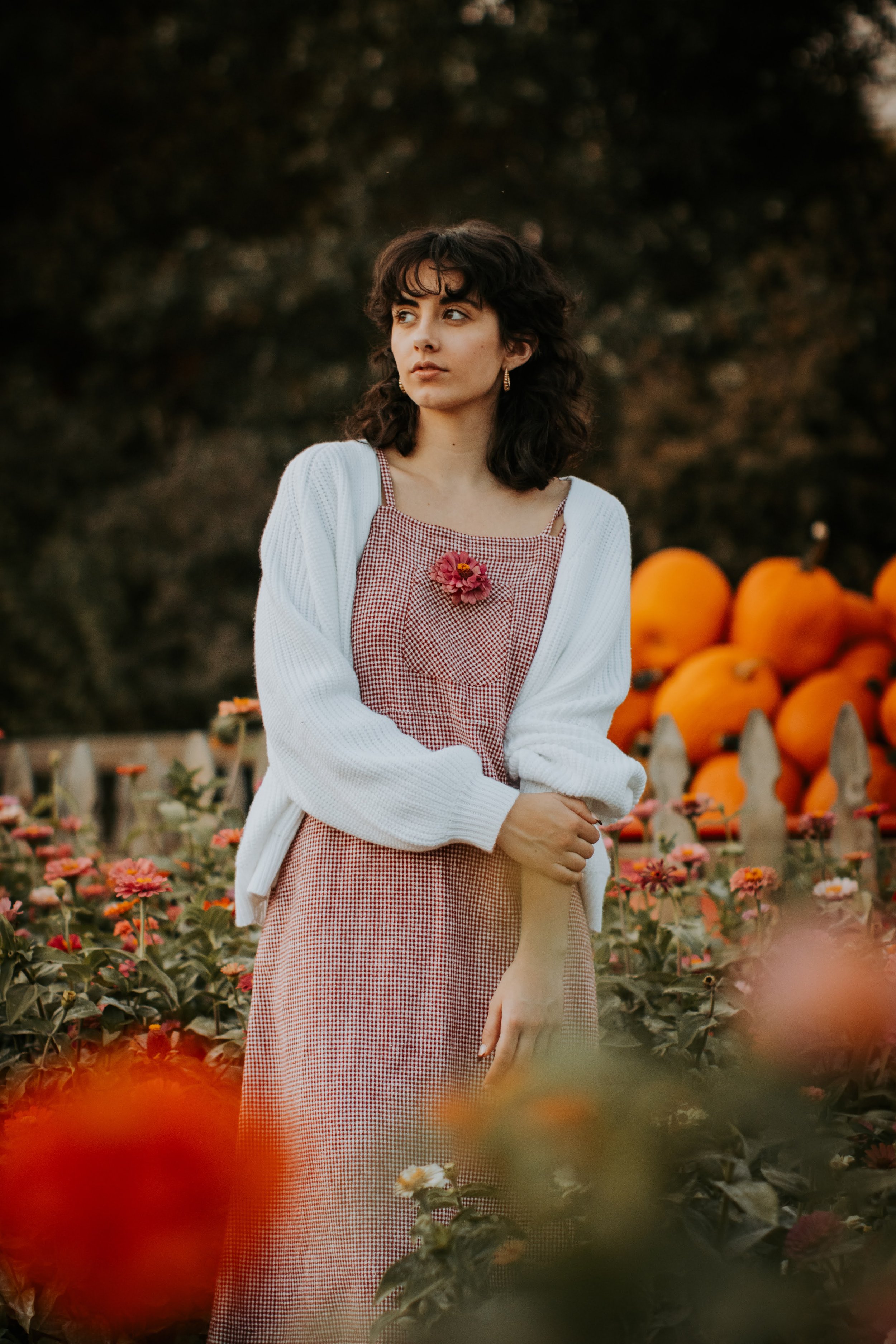 A young woman standing in a pumpkin patch surrounded by orange pumpkins and flowers, wearing a red and white checkered dress with a white cardigan, and a pink flower pin on her chest.