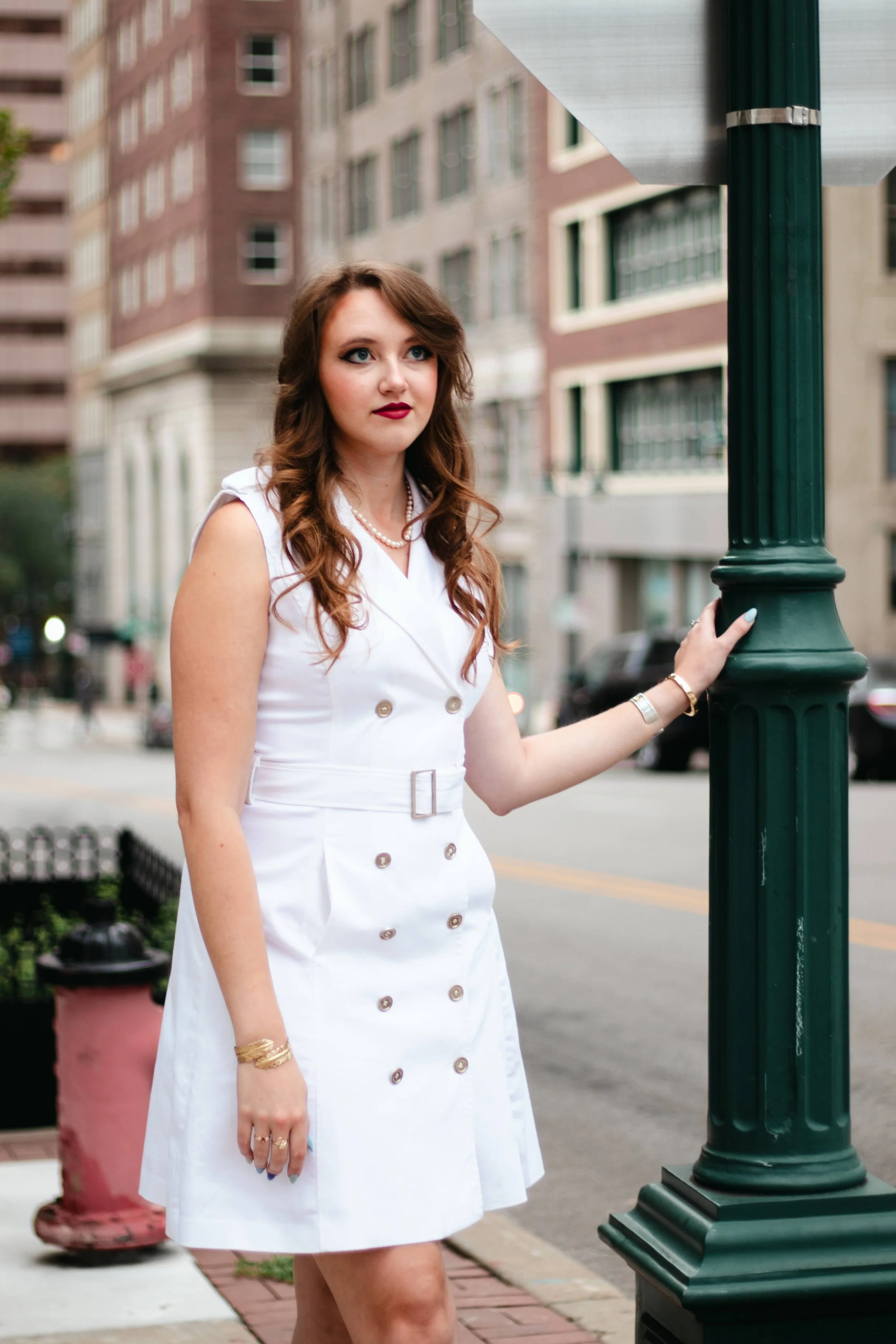 A woman with long, wavy brown hair and red lipstick, wearing a white sleeveless dress with gold buttons, standing on a city sidewalk holding onto a green lamppost, with tall buildings in the background.