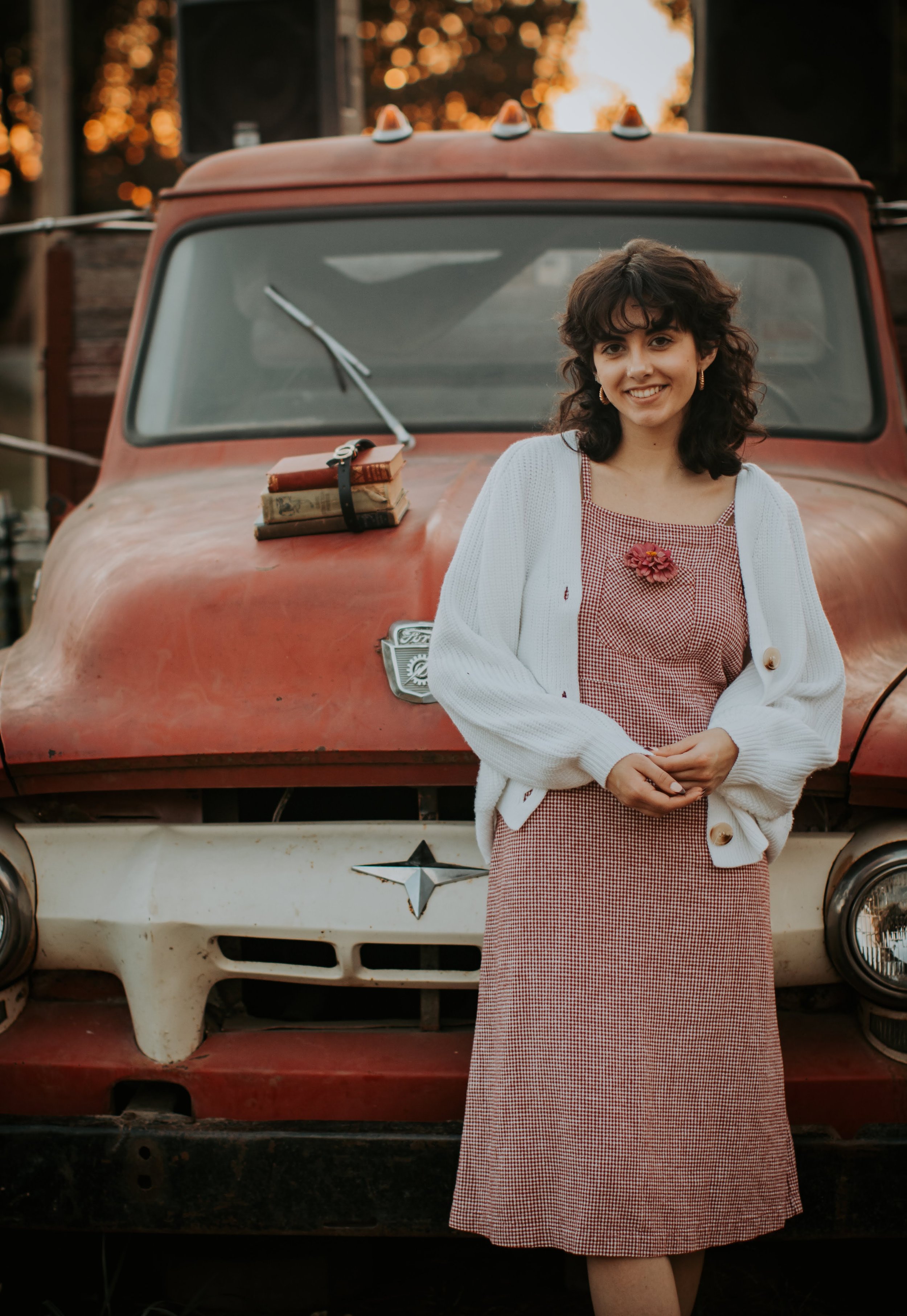 A young woman with curly dark hair and a white cardigan standing in front of a vintage red truck with a stack of old books on the hood, during sunset.
