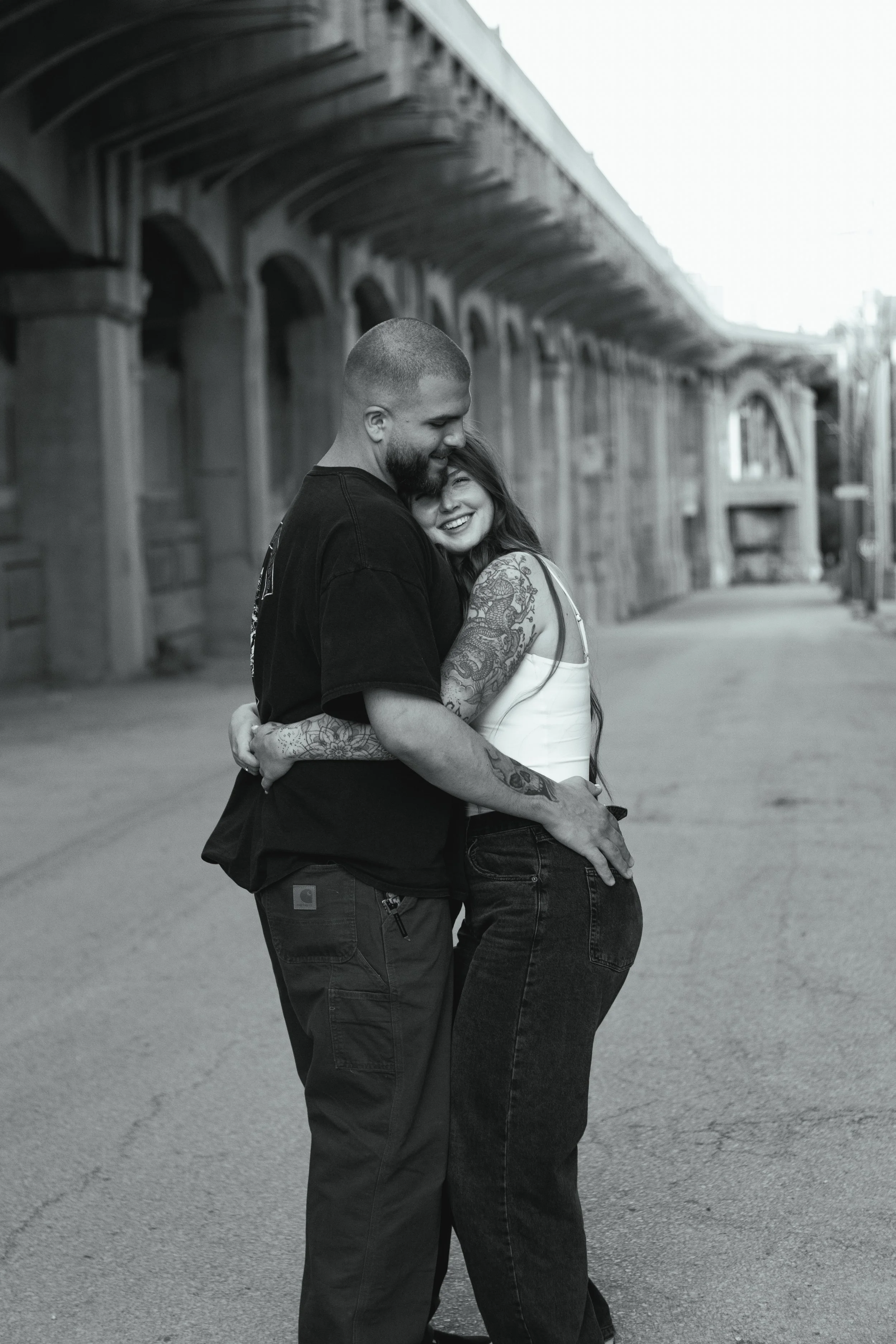 A black and white photo of a smiling couple hugging on an empty street under a bridge.
