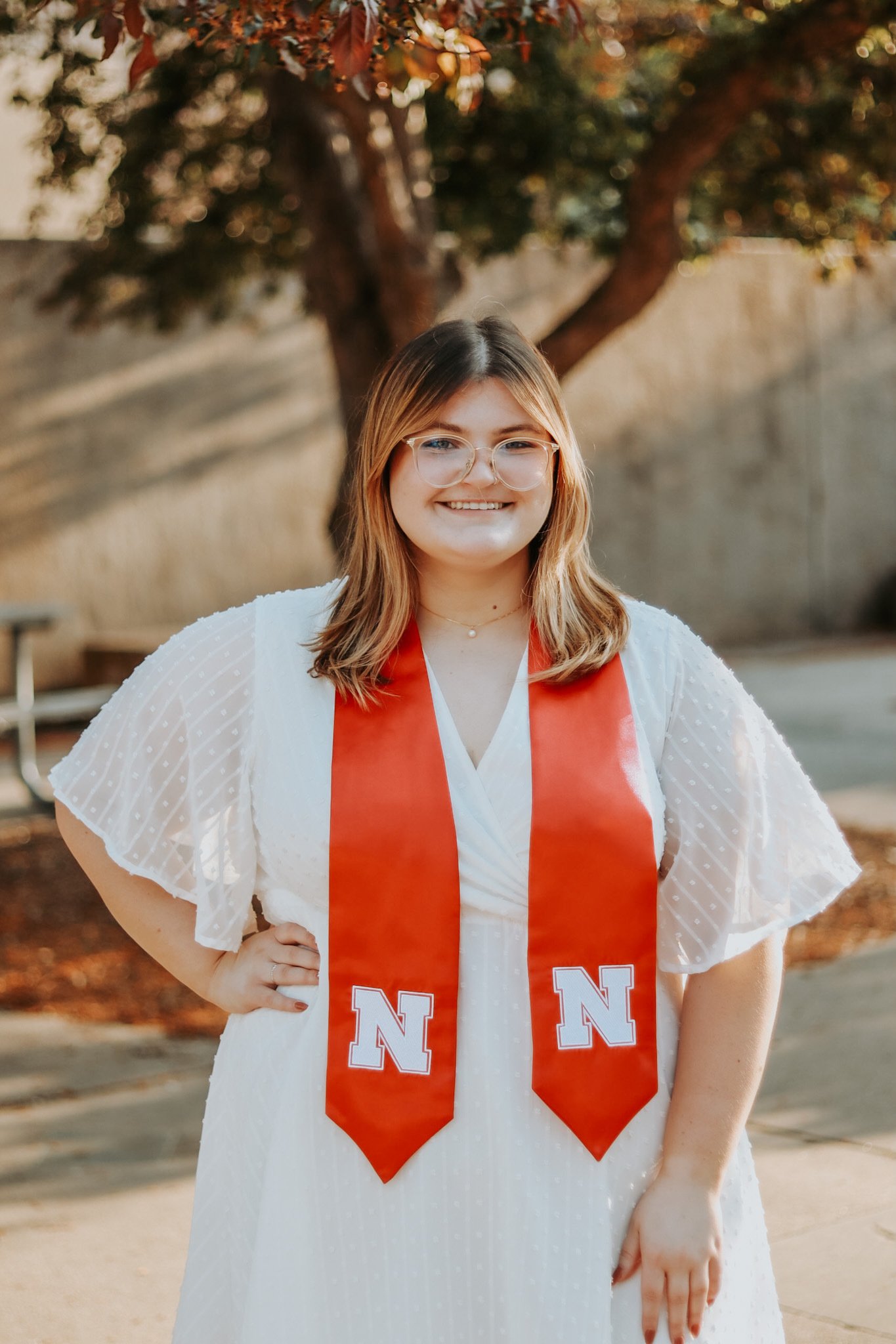 A young woman with glasses, wearing a white dress and an orange graduation stole with a white letter 'N' on each end, stands outdoors with hands on hips, smiling at the camera. There are trees and a concrete wall in the background.