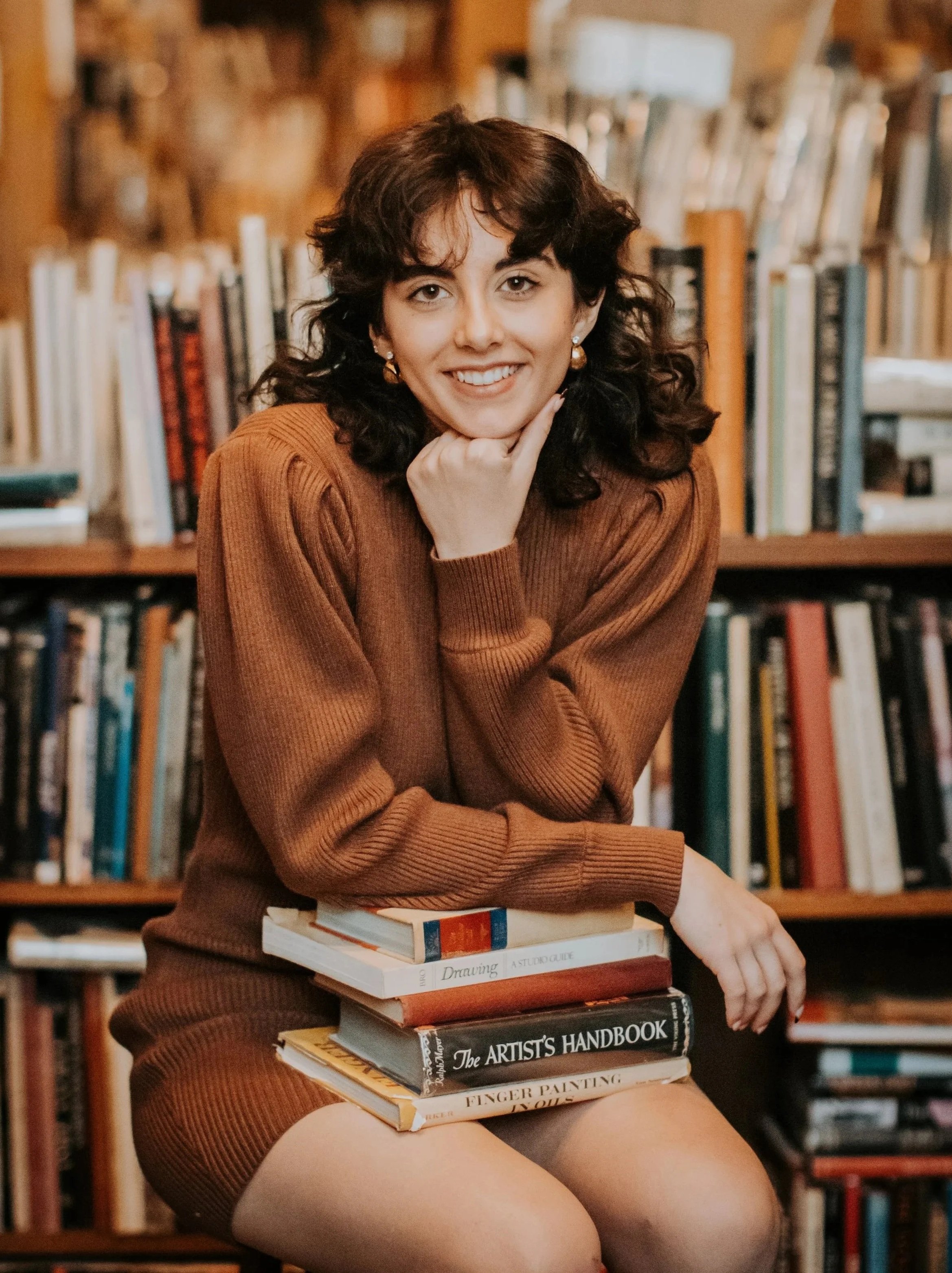 A young woman with dark wavy hair, wearing a brown sweater, smiling and sitting in a library, resting her chin on her hand, with books stacked on her lap and more books on shelves behind her.