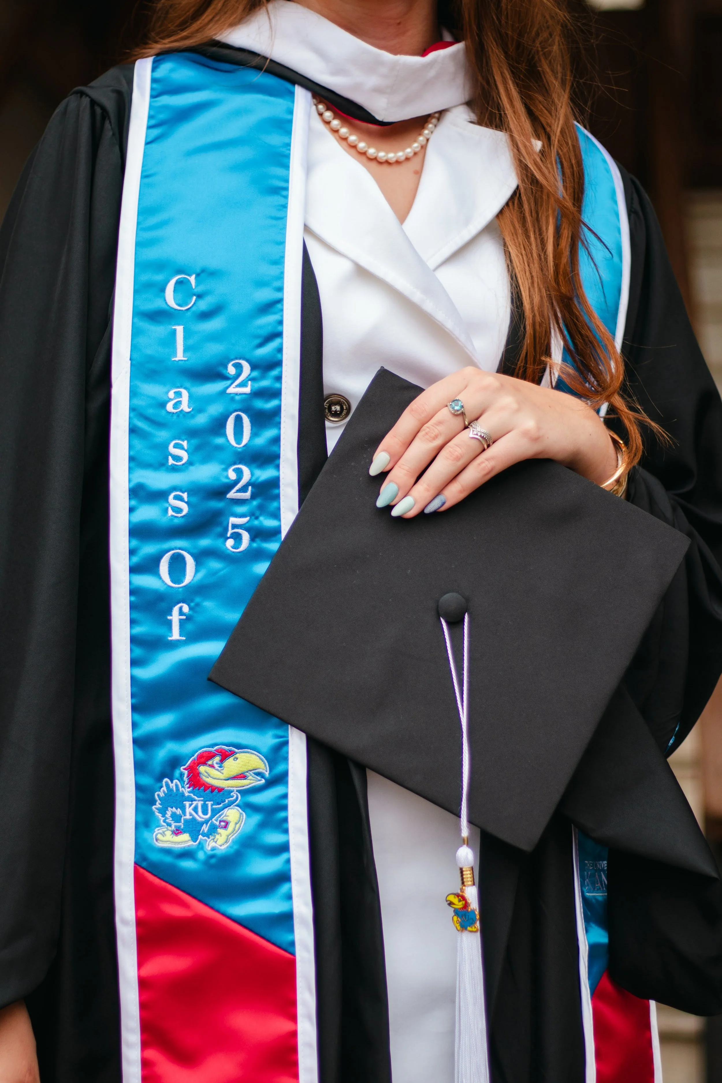 Close-up of a graduate wearing a black cap and gown, with a blue and white stole that has 'Class of 2025' and the Kansas Jayhawks logo. The graduate is holding a black cap in front of their chest, with painted nails, rings, and a pearl necklace visible.