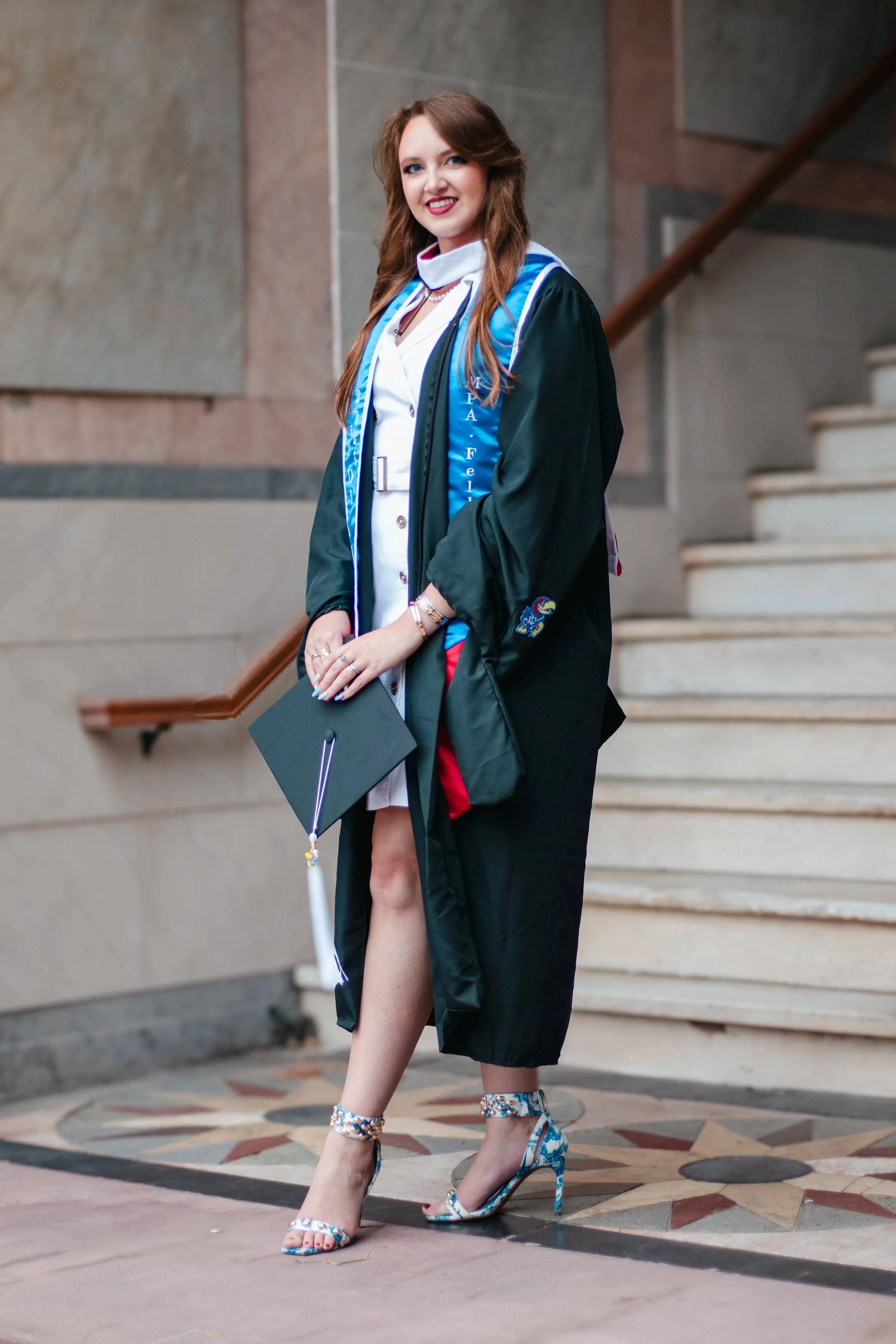 Woman in graduation gown and cap holding diploma, posing on stairs indoors.