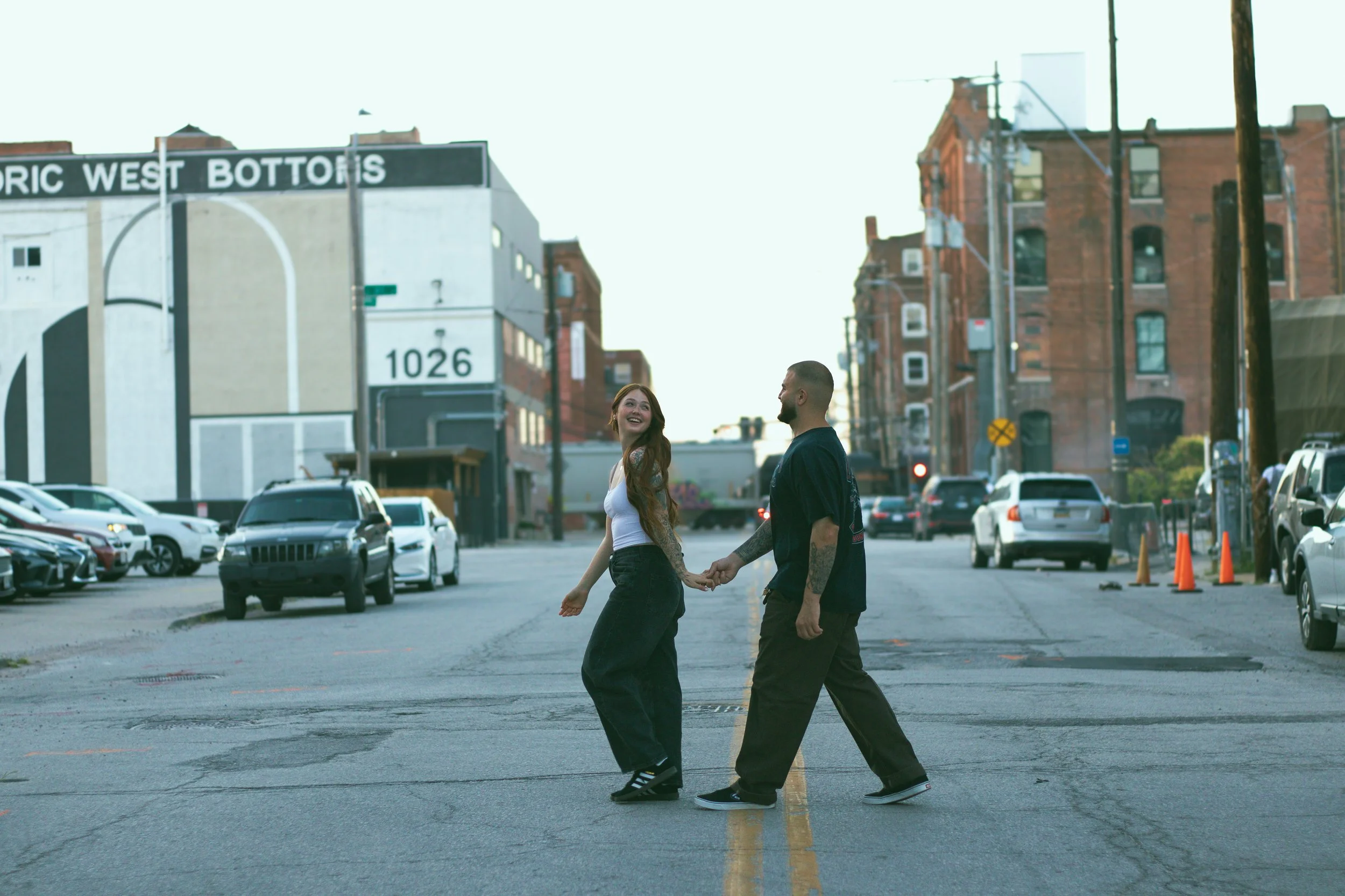A young woman and a young man holding hands and smiling at each other while crossing a city street. The street is lined with parked cars and brick buildings in the background.