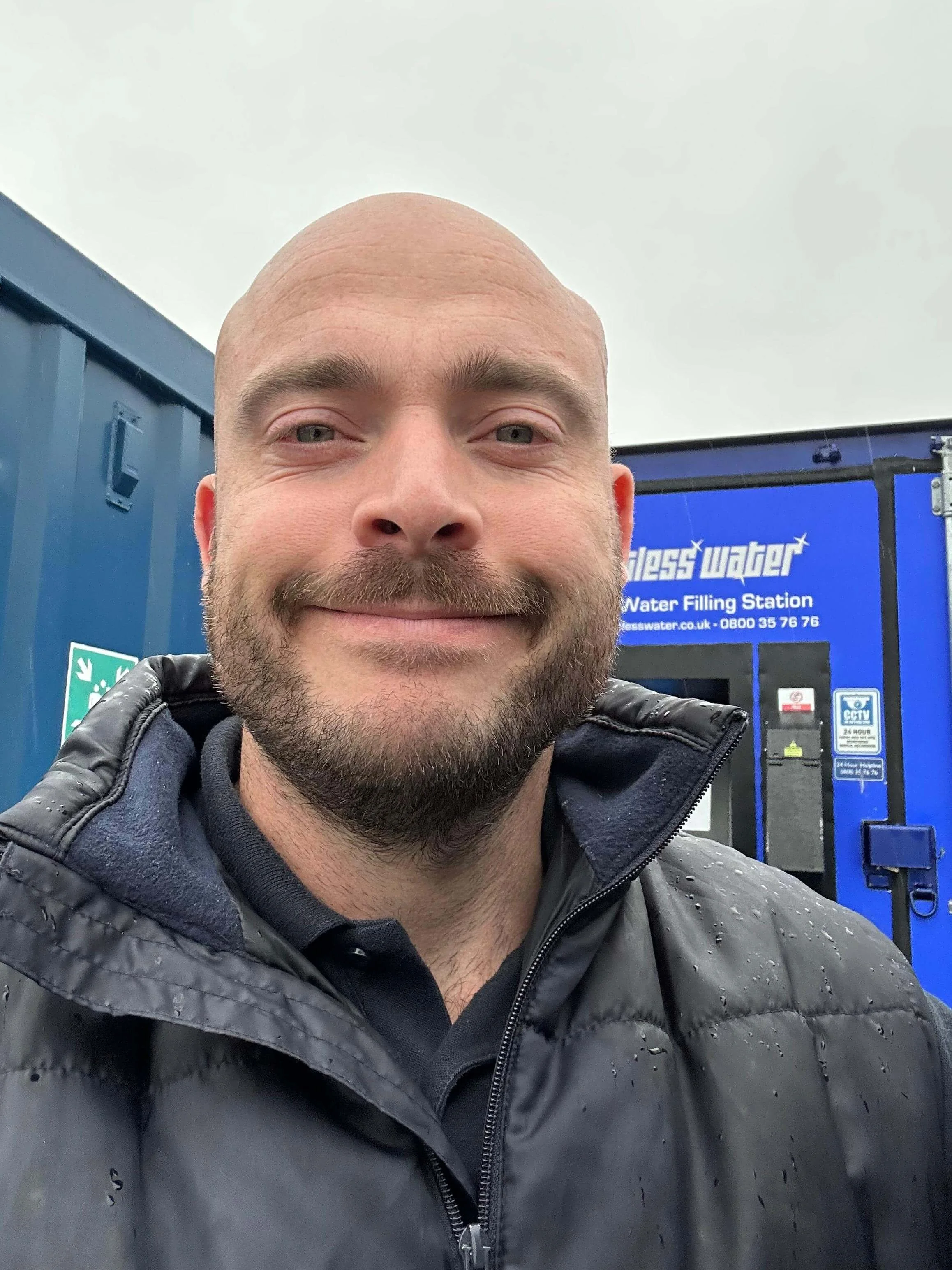 A smiling man taking a selfie outdoors, standing in front of a blue water filling station.
