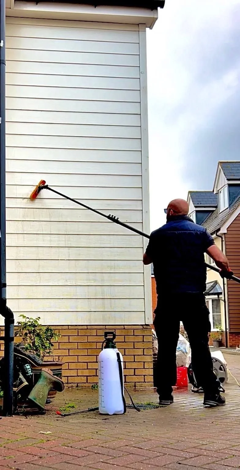 A man cleaning the exterior wall of a house using a long-handled cleaning tool. The house has white siding and a brick foundation, with a garden hose and a white water container nearby. Other houses are visible in the background.