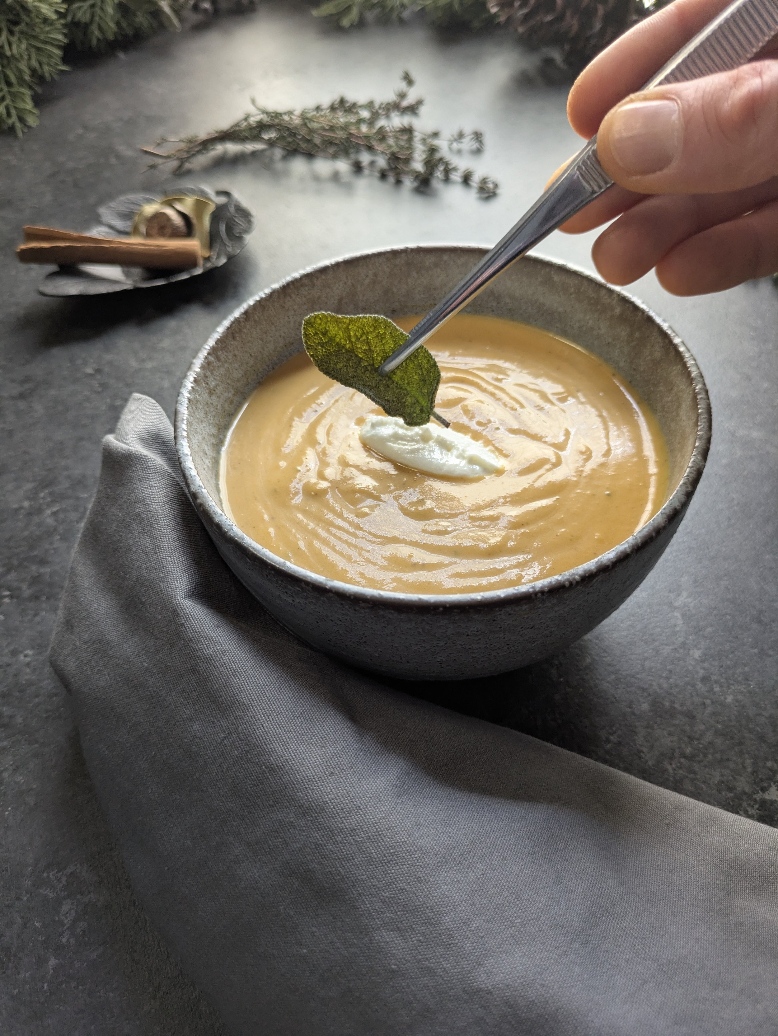 butternut bisque served in an earthenware bowl with a dollop of sour cream in the middle, being garnished by a hand holding forceps with a fried sage leaf.