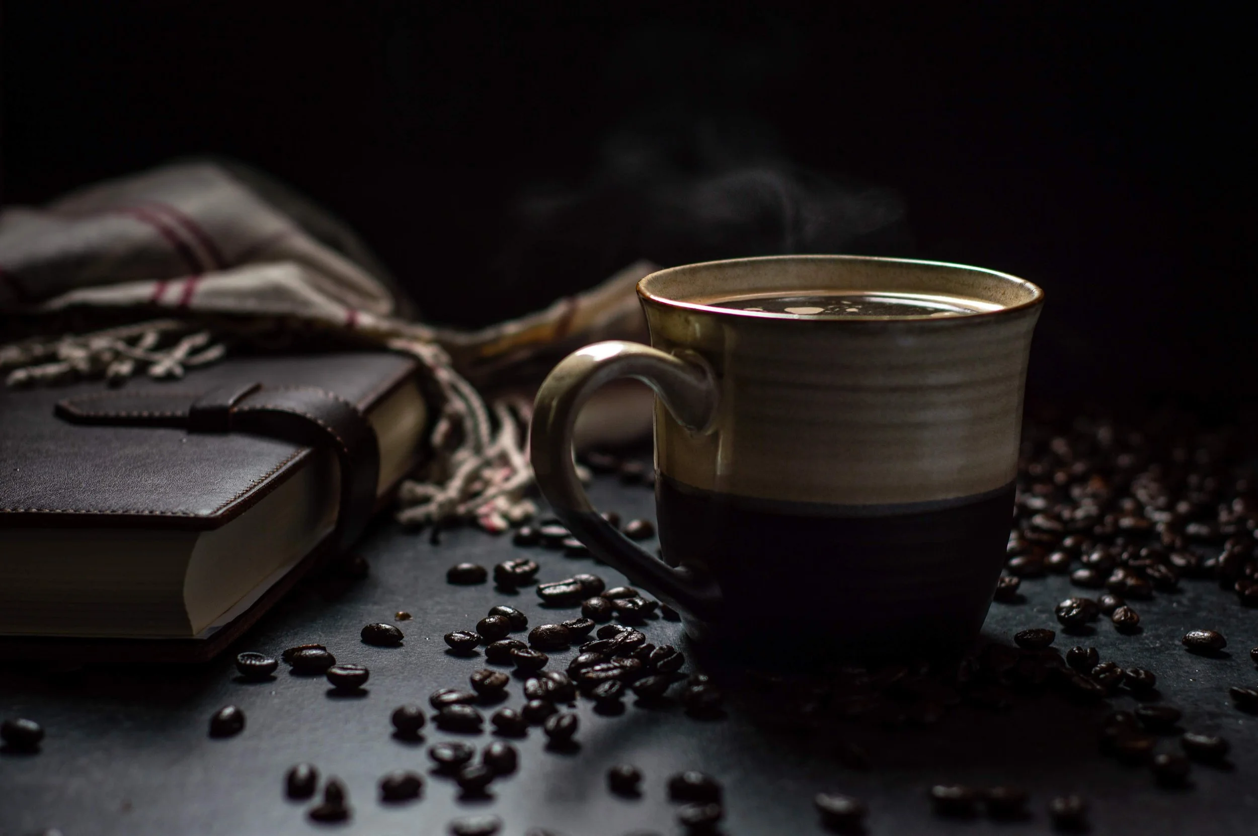 A cup of steaming hot coffee next to a leather book and a scarf and scattered coffee beans