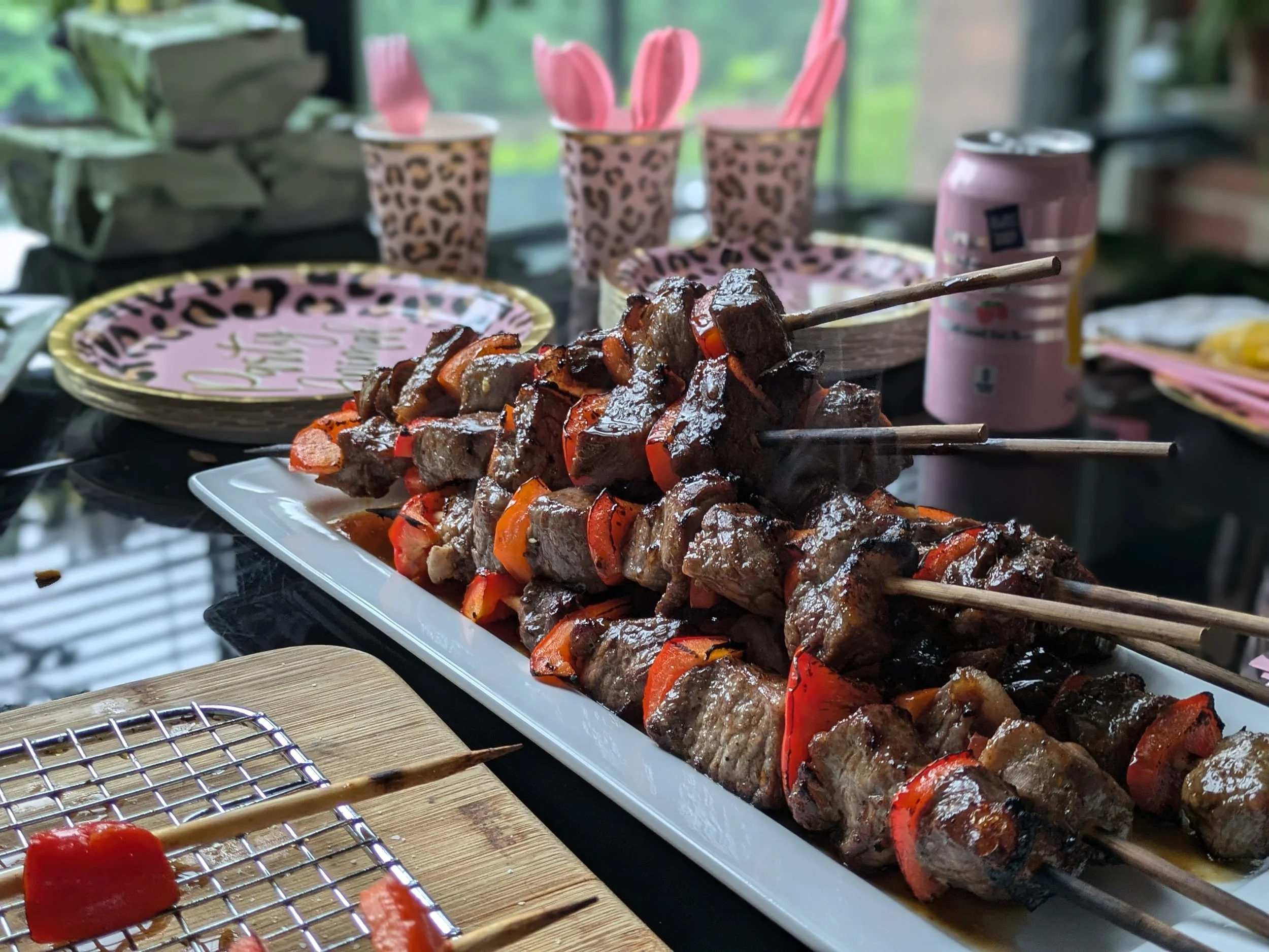 Close-up of skewered grilled meat and vegetables on a white platter, with a small grilled piece and a skewer on a wooden board in the foreground. In the background, there are pink napkins, leopard print cups, and a can of soda, with a pink tray that 