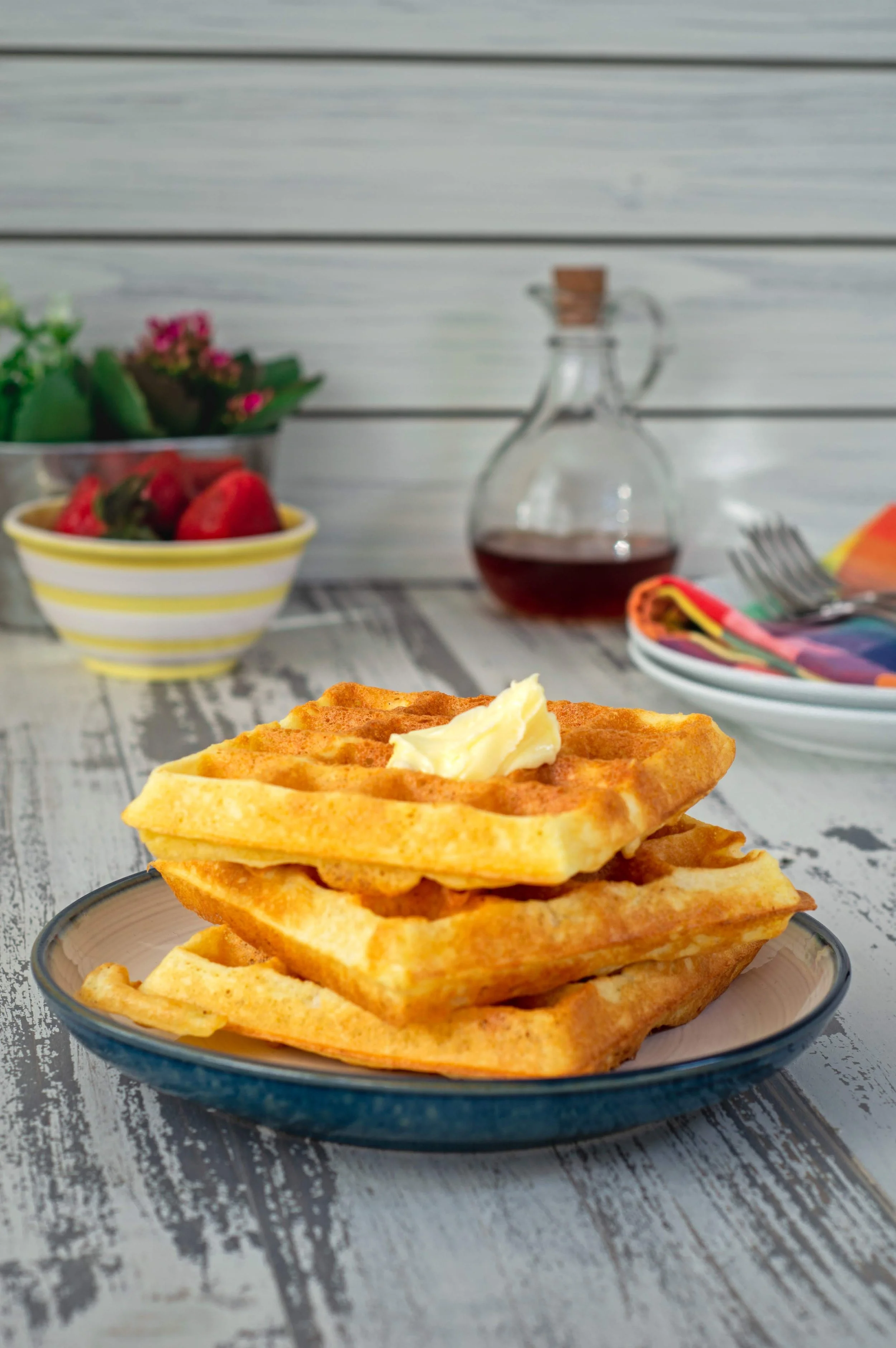 A stack of waffles with butter, maple syrup, and a bowl of fresh strawberries