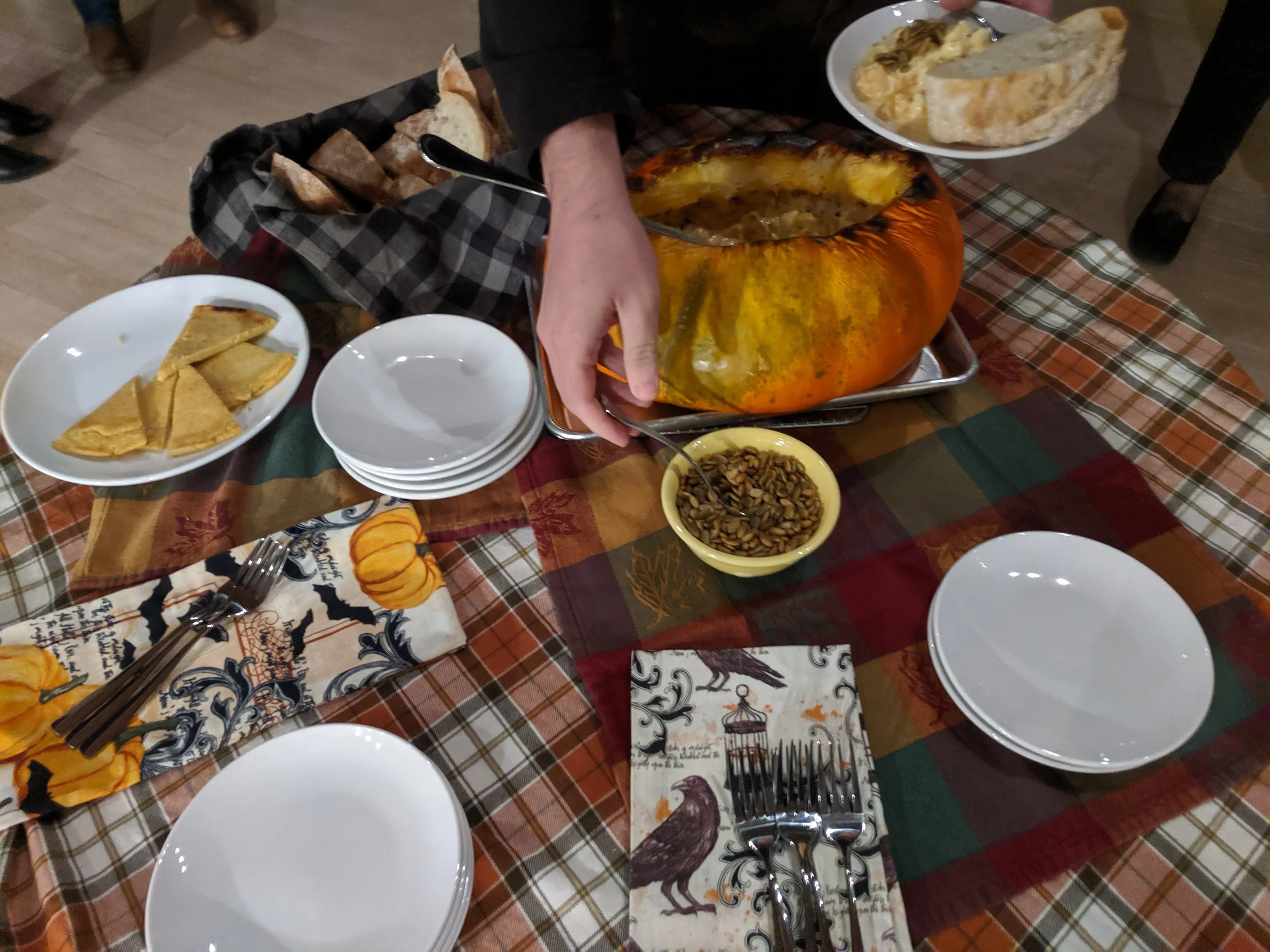 A baked stuffed pumpkin station with pumpkin fondue, bread, socca, pepitas. A guest spoons pepitas on their dish.